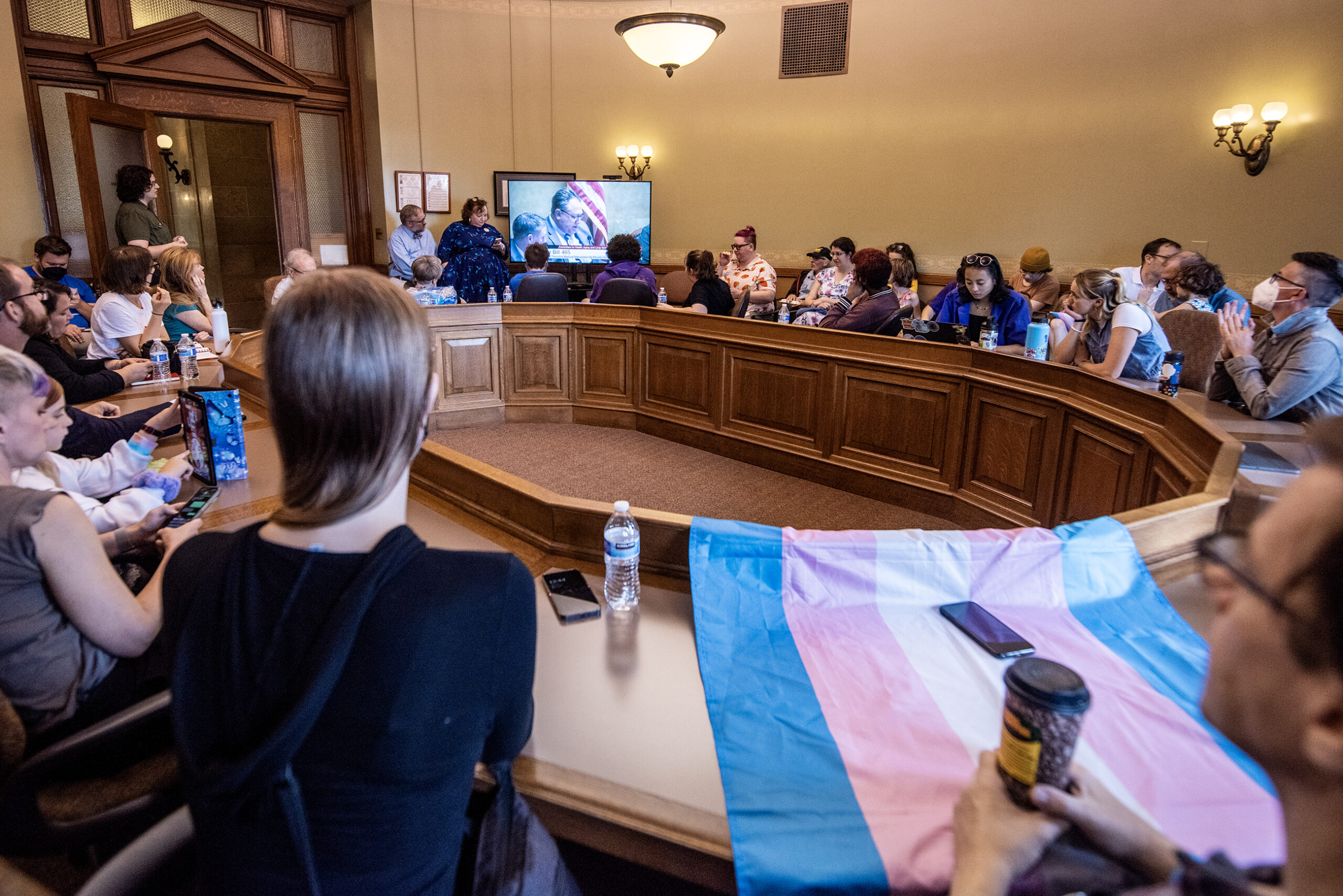 A trans flag is draped over a table as people watch the hearing on tv.