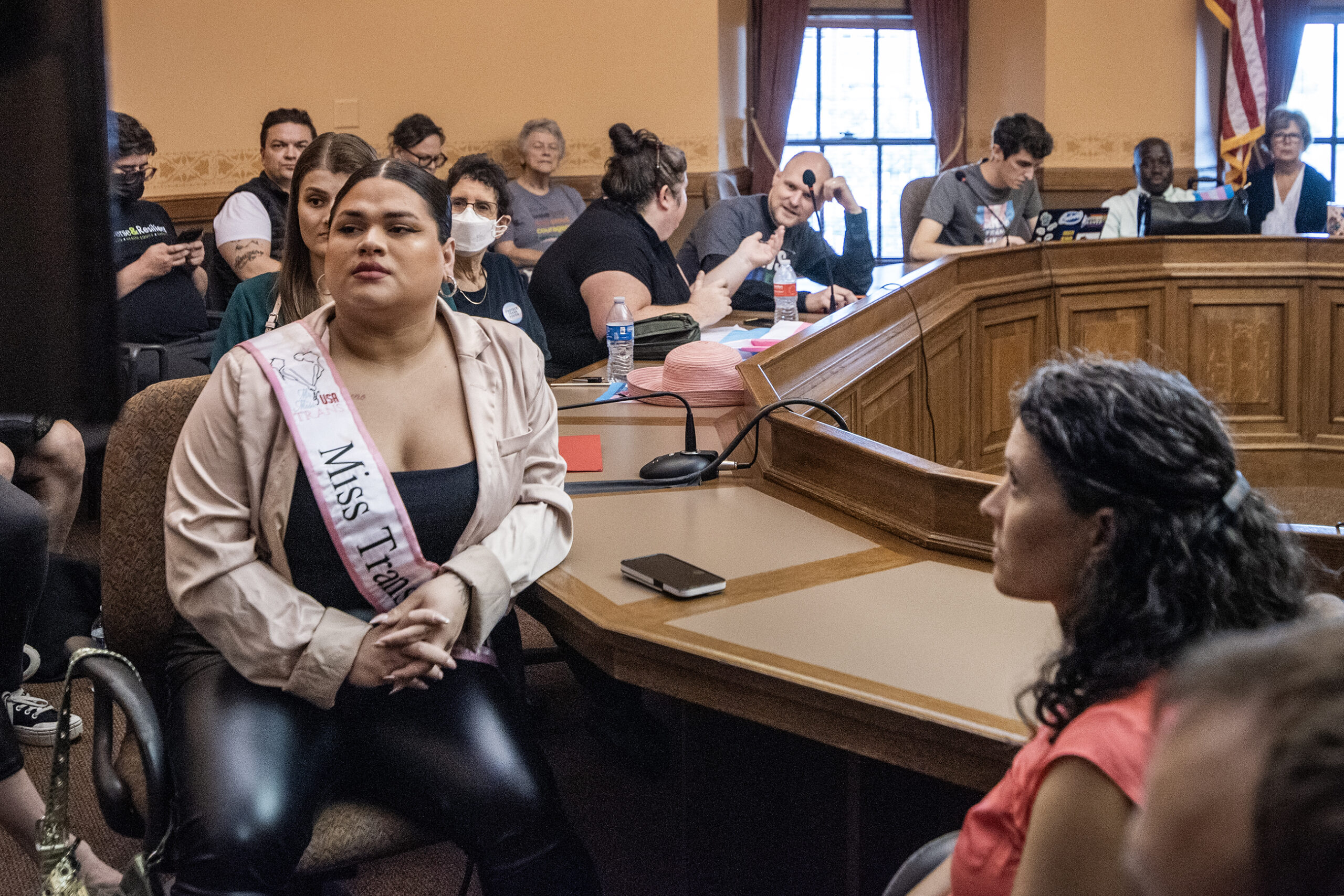 A person in a sash watches the hearing on tv with a room of people.