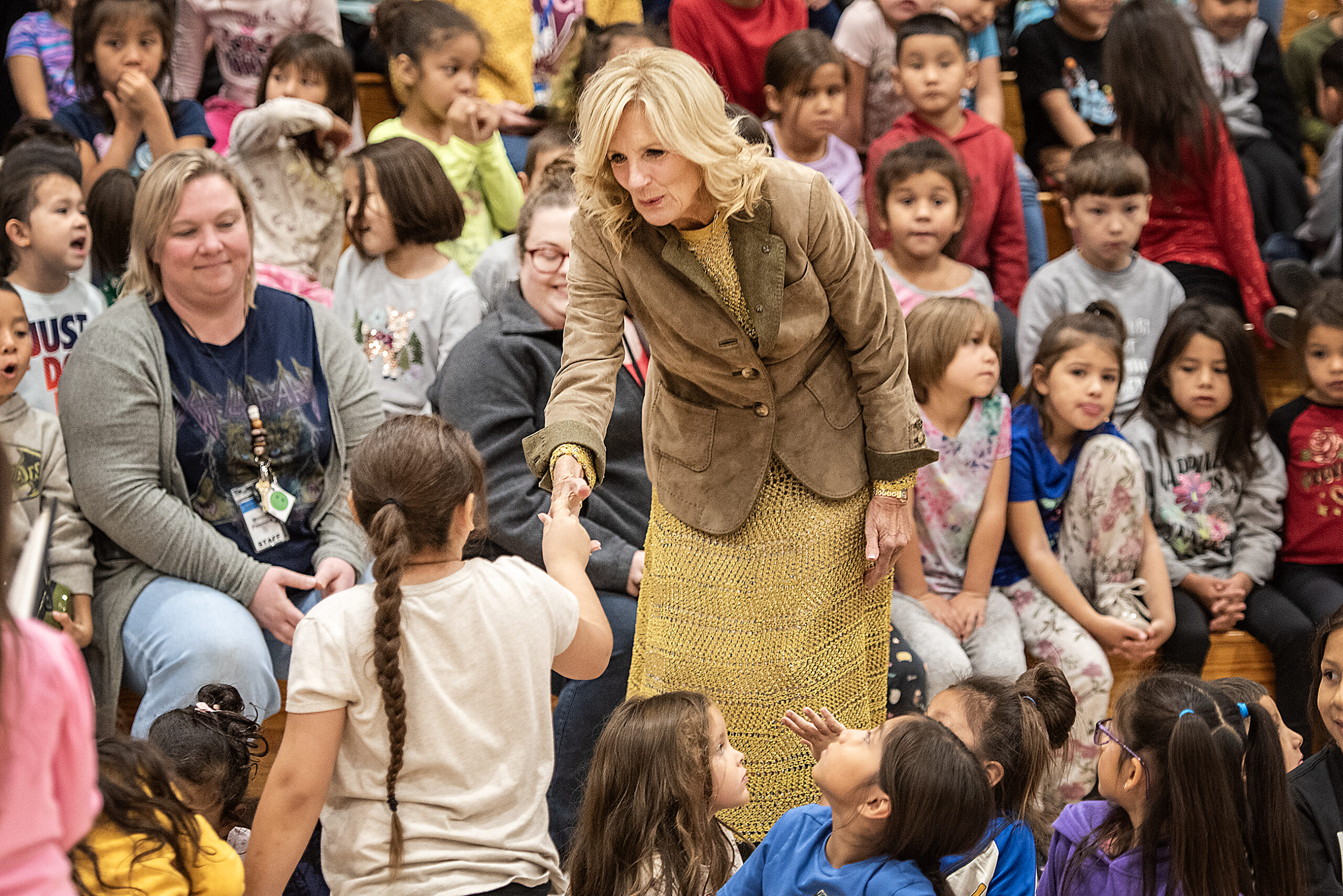Jill Biden smiles as she leans over to shake the hand of a small girl in a crowded gym of excited children.