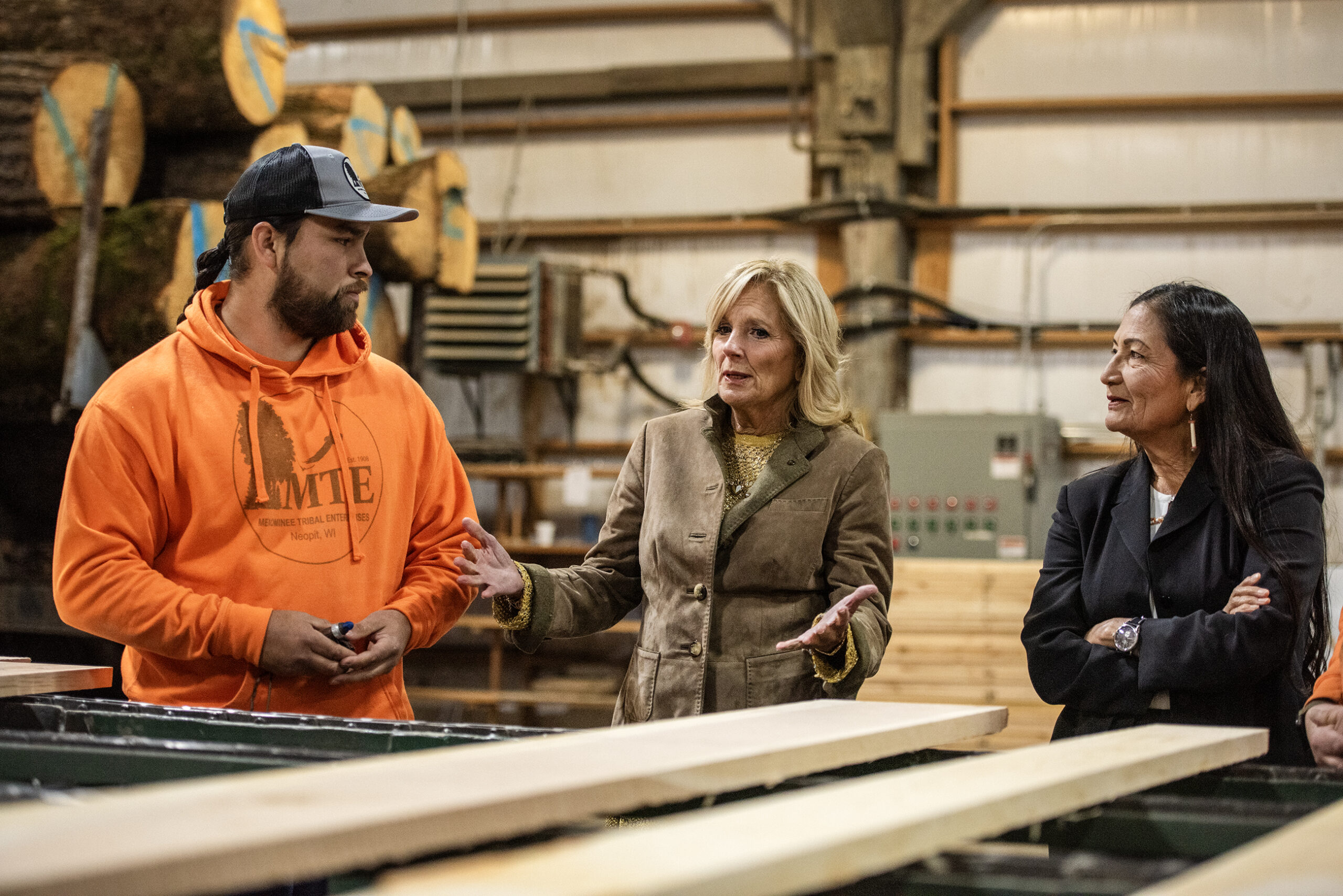 Jill Biden and Deb Haaland stand in front of wooden planks as a worker answers their questions.