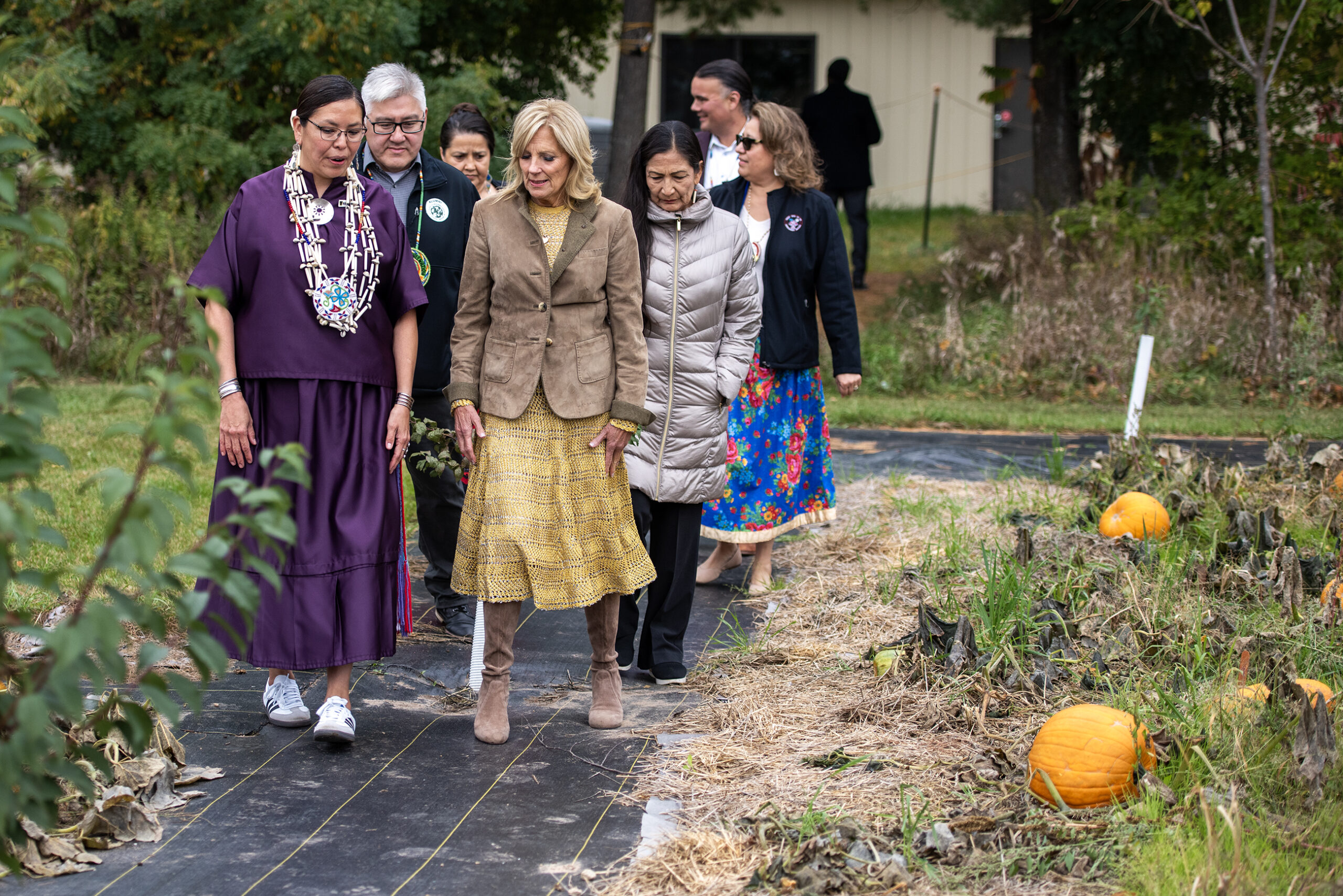 Jill Biden and Deb Harland walk with other leaders through a garden. Pumpkins can be seen growing in a patch.