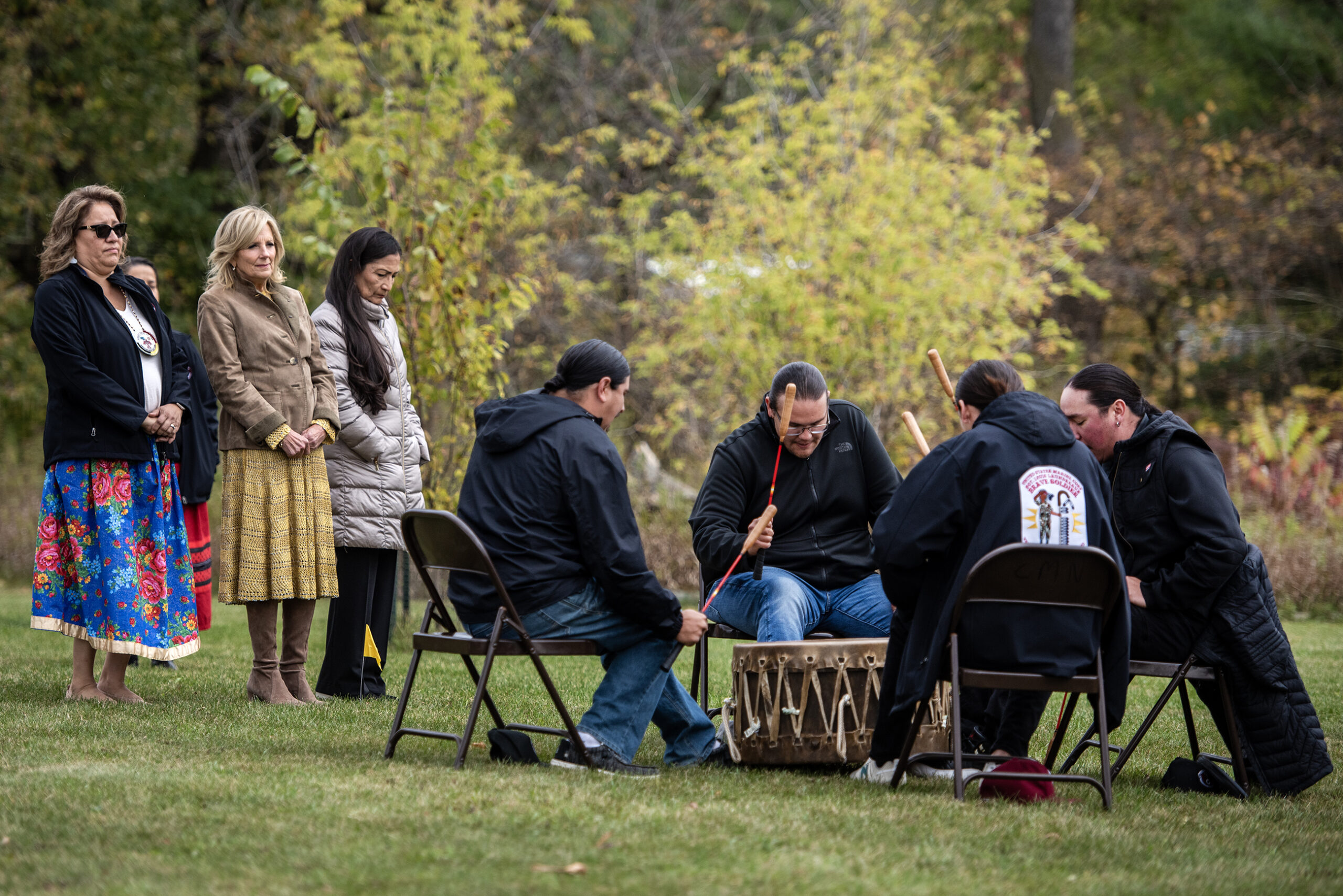 Four men drum and sing outside as people watch.