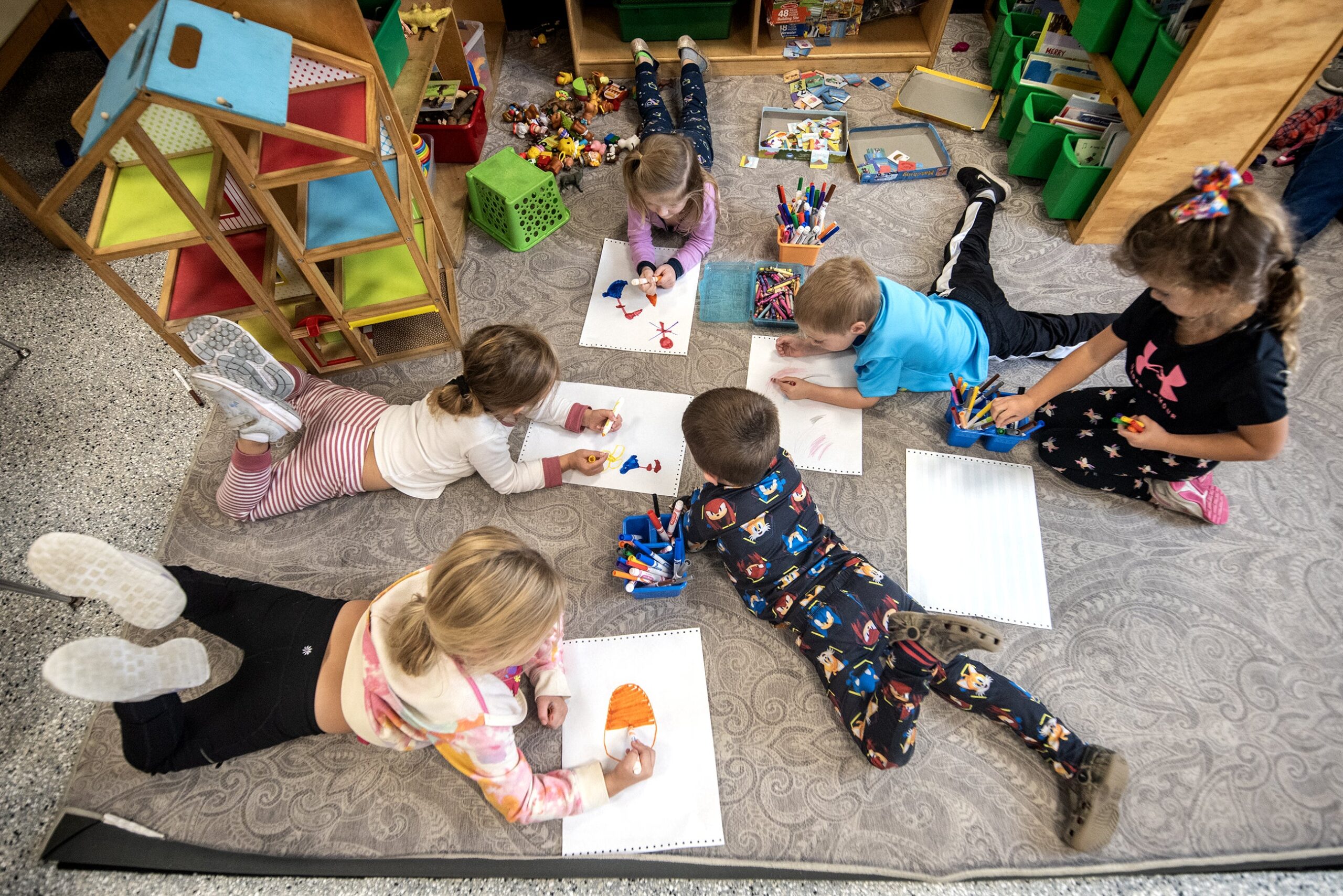 Six students are seen from above as they lay on the floor as they draw pictures with markers.