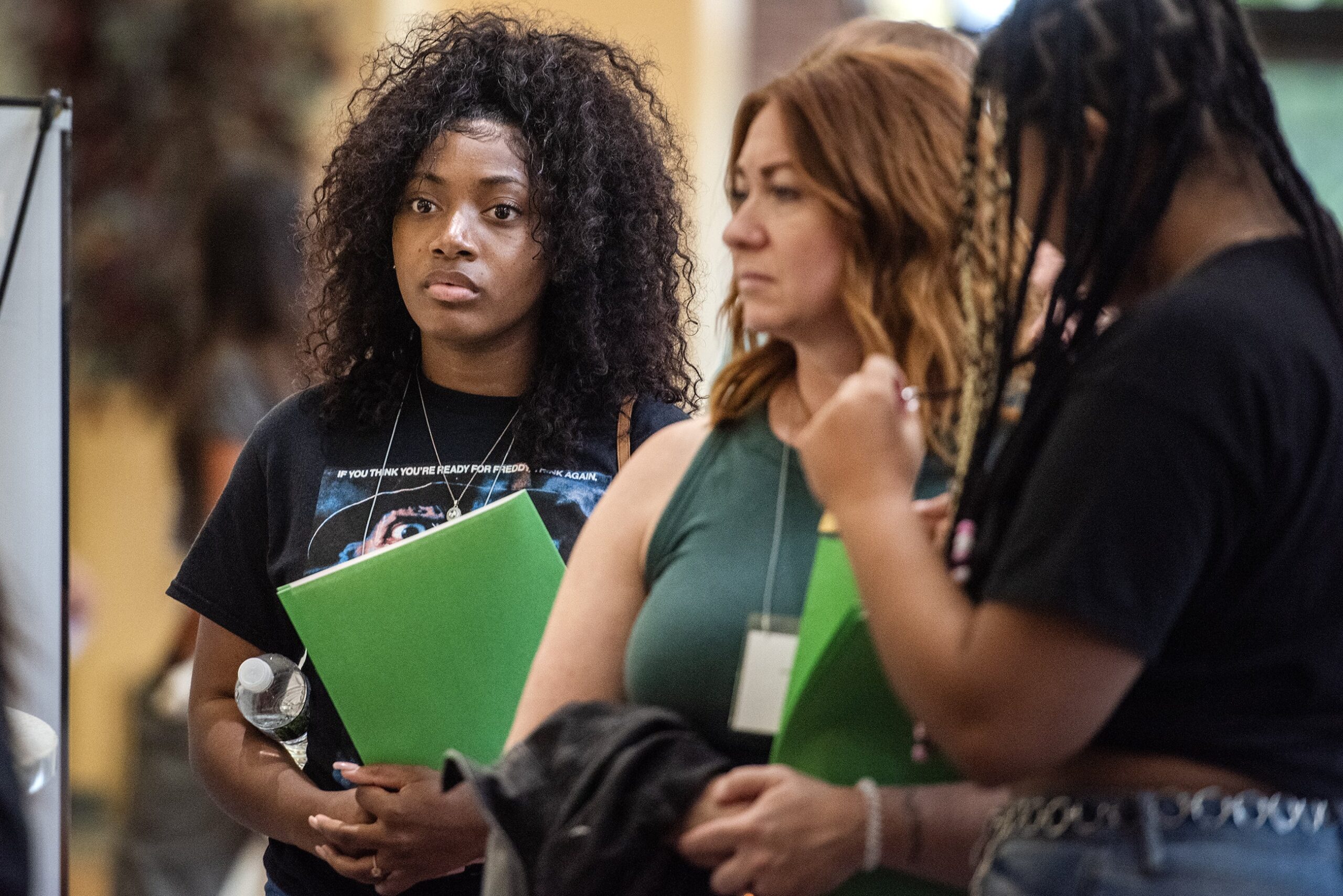 Three students hold folders as they wait in line.