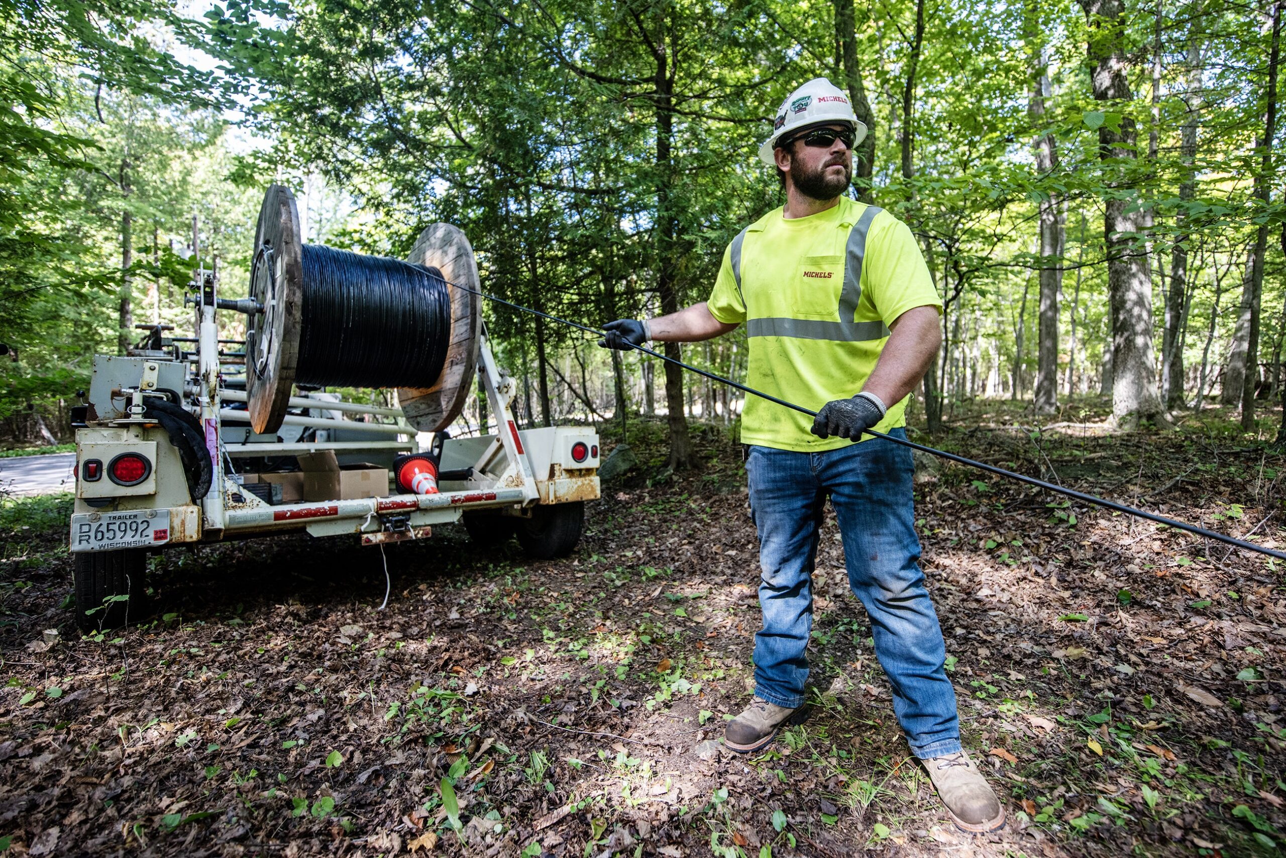 A worker in neon yellow stands in a wooded area as he pulls a black cable from a large spool.