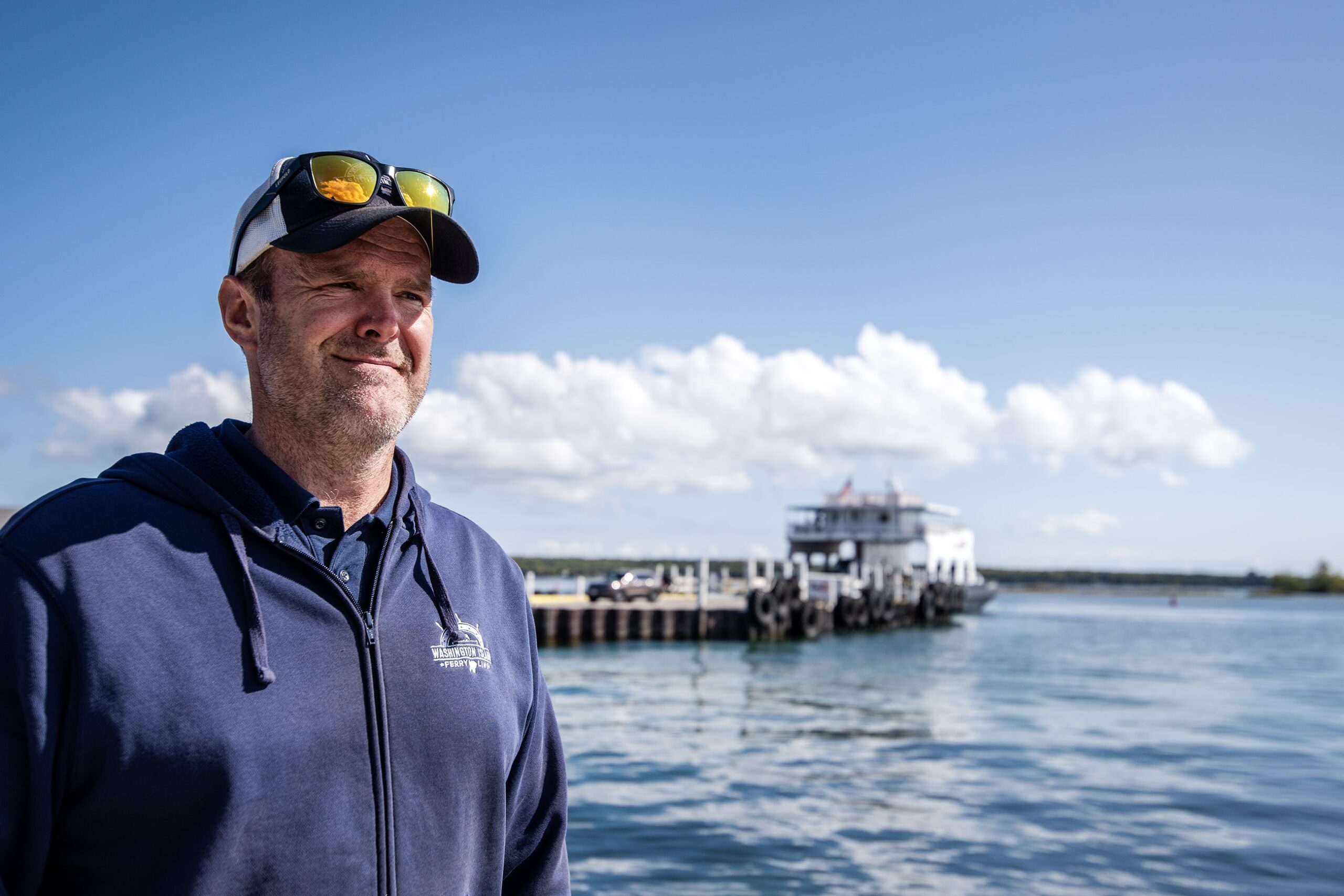 A man stands near the shore with a blue sky behind him as a ferry boat approaches.