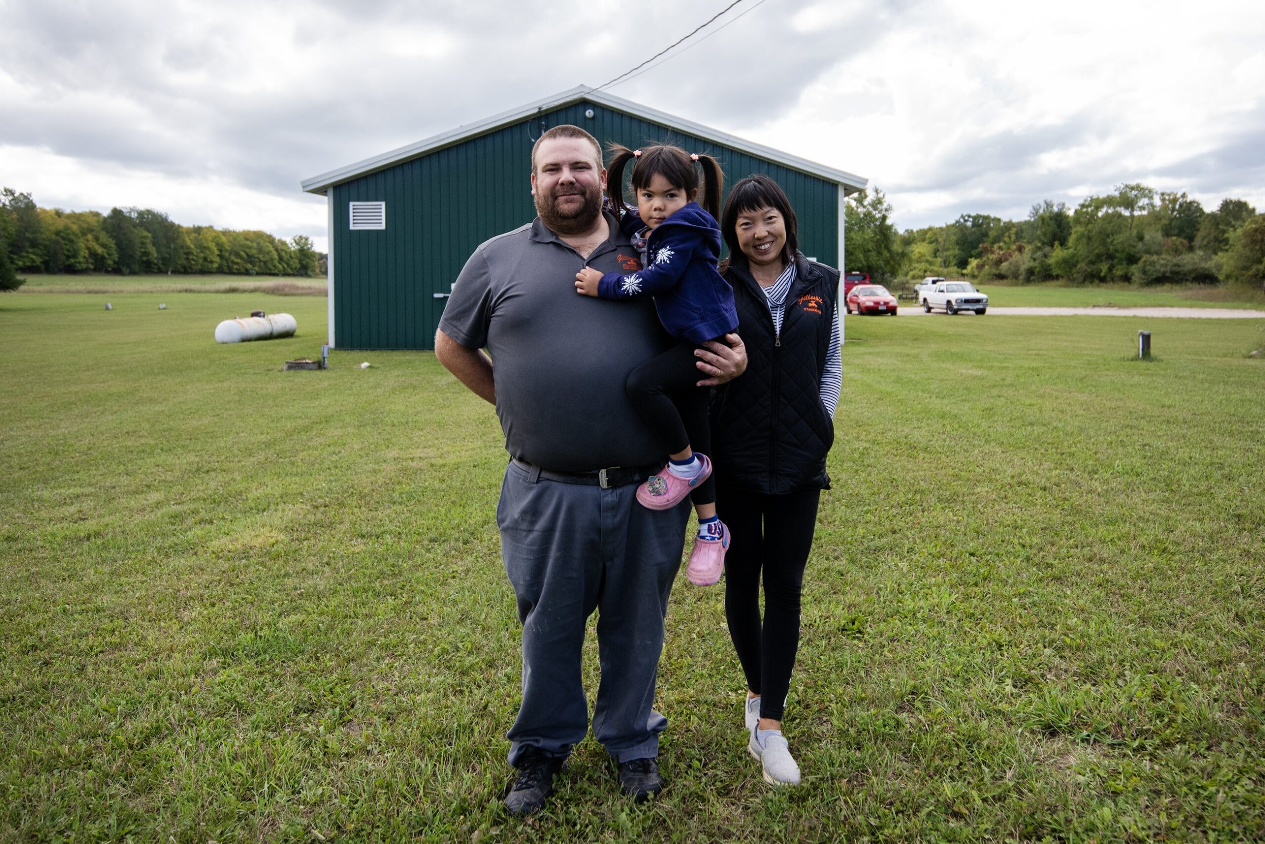 A family of three stands in a grassy lawn outside of a blue office building.