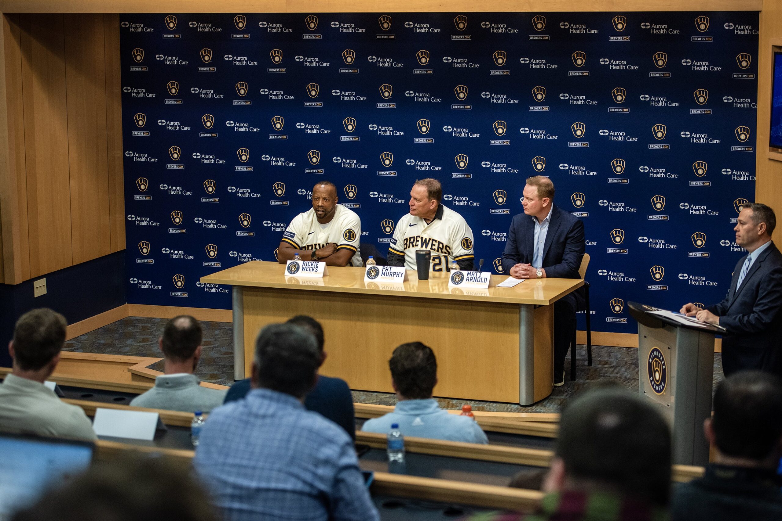 The three men sit at a wooden table as members of the press attend.