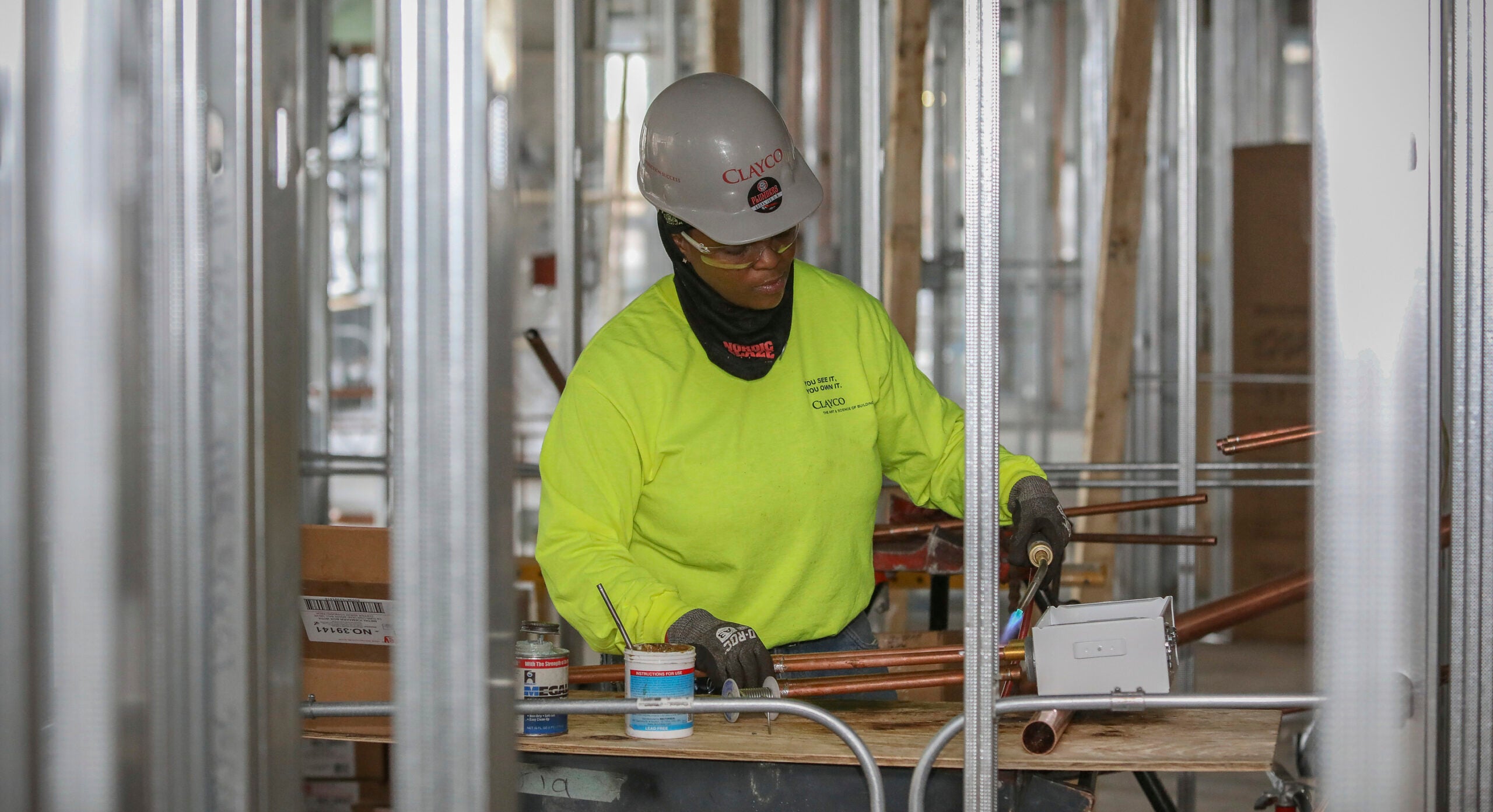 A woman dressed in a hard hat and high visability gear welds some copper pipes.
