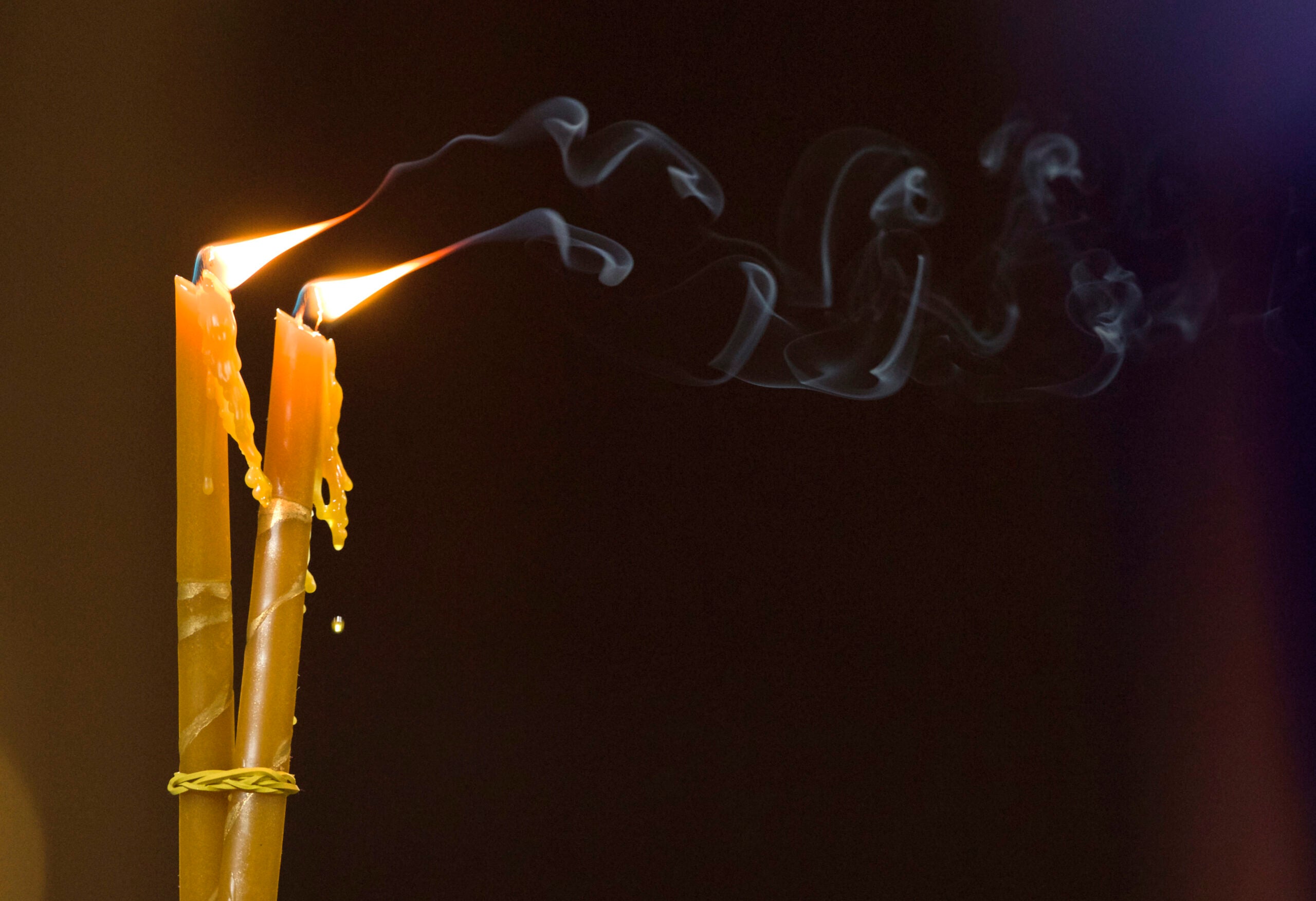 Two long, thin candles are buring side by side. Soot or smoke from the candles drifts away from them.