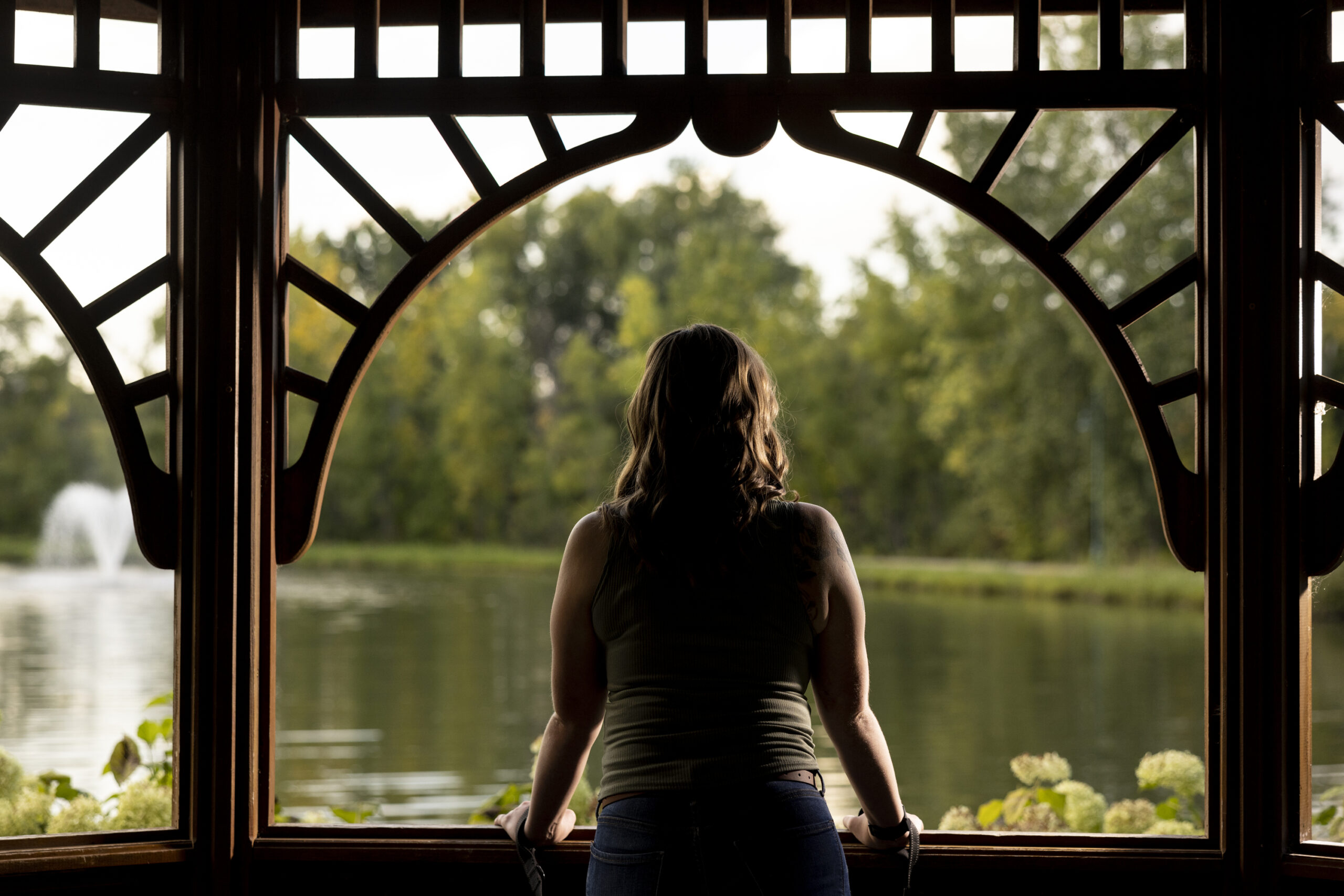 A woman stands with her back to the camera, in a gazebo, facing a body of water