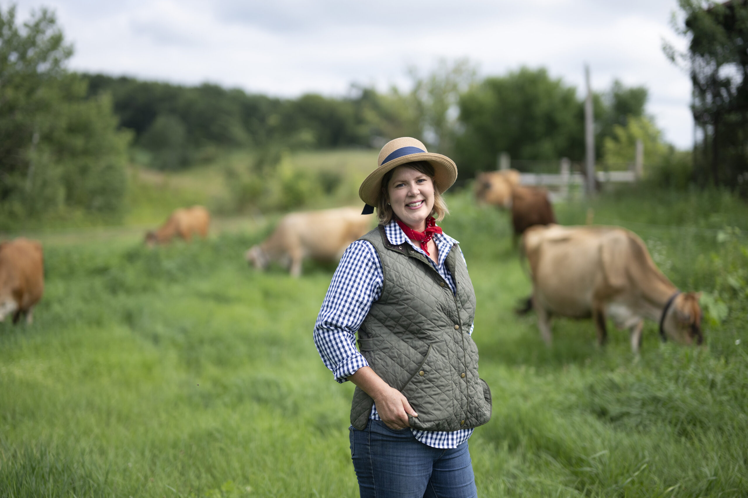 A white woman wearing a hat stands in front of brown cows grazing in a field.