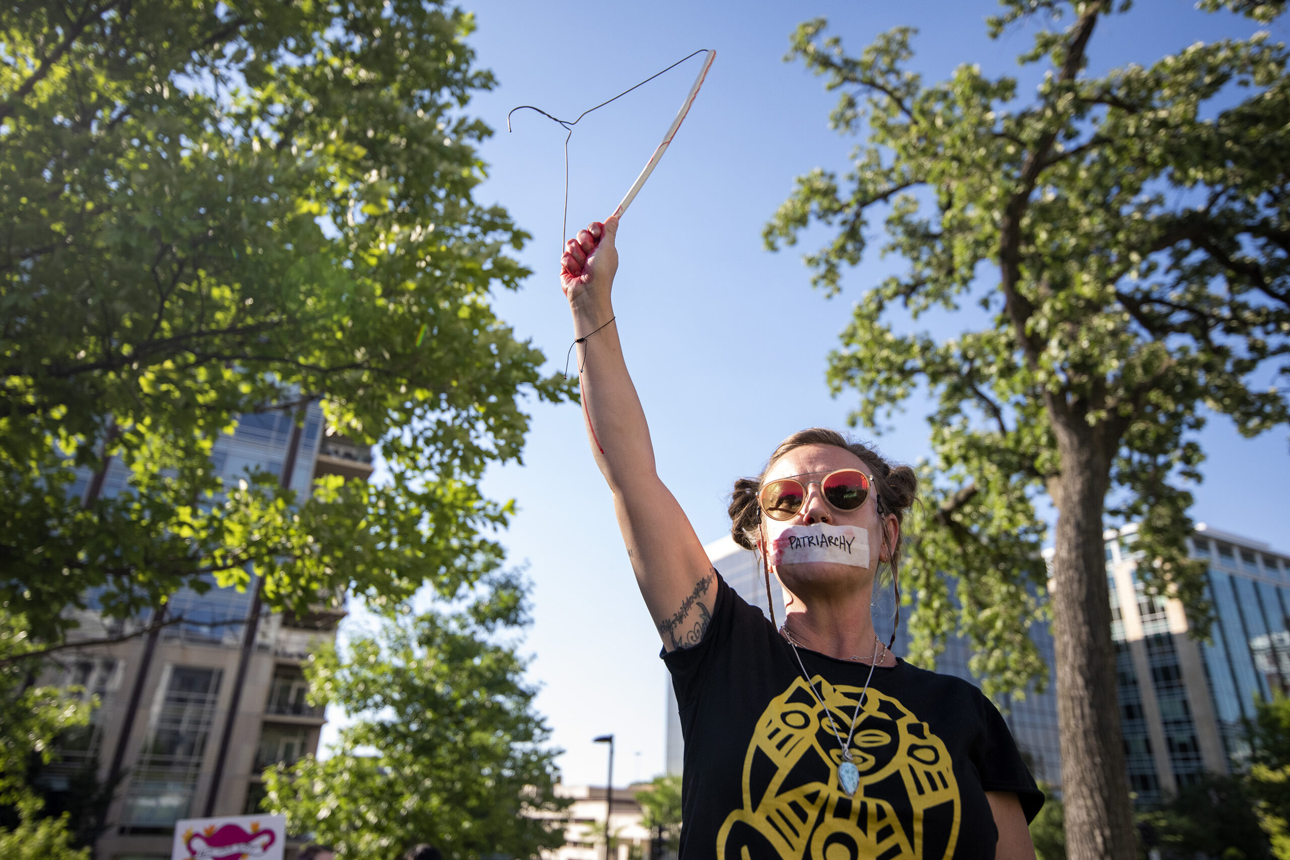 A protester has a piece of tape over her mouth that says "patriarchy." She holds a hanger in the air.