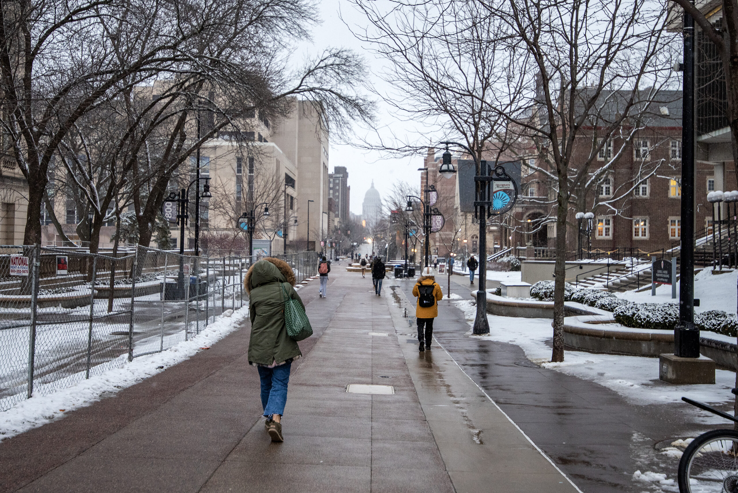 Students walk outside on a sidewalk while wearing coats. The Wisconsin State Capitol can be seen in the background.