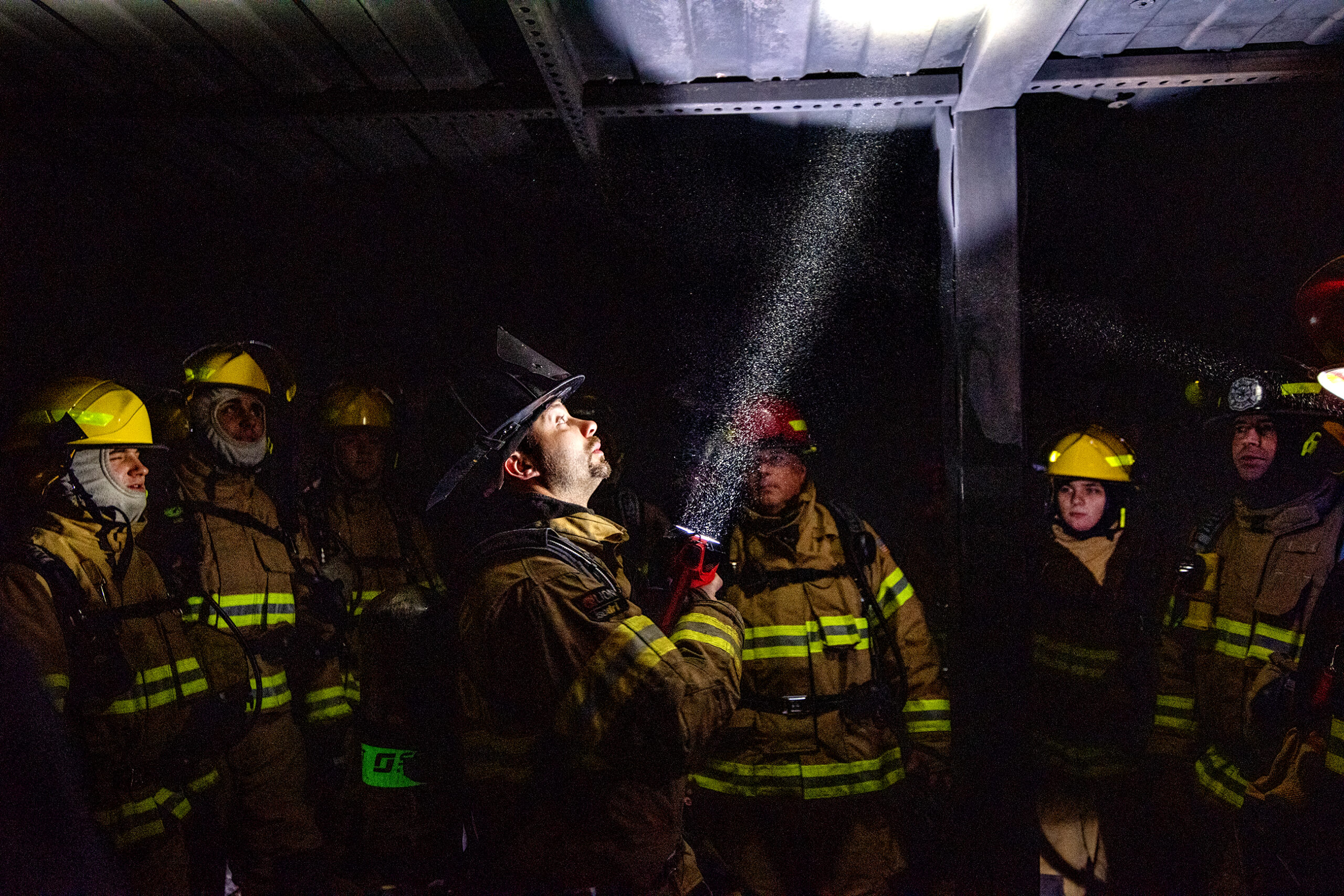 A man in a firefighter uniform shines a flashlight into a dark room as students gather around.