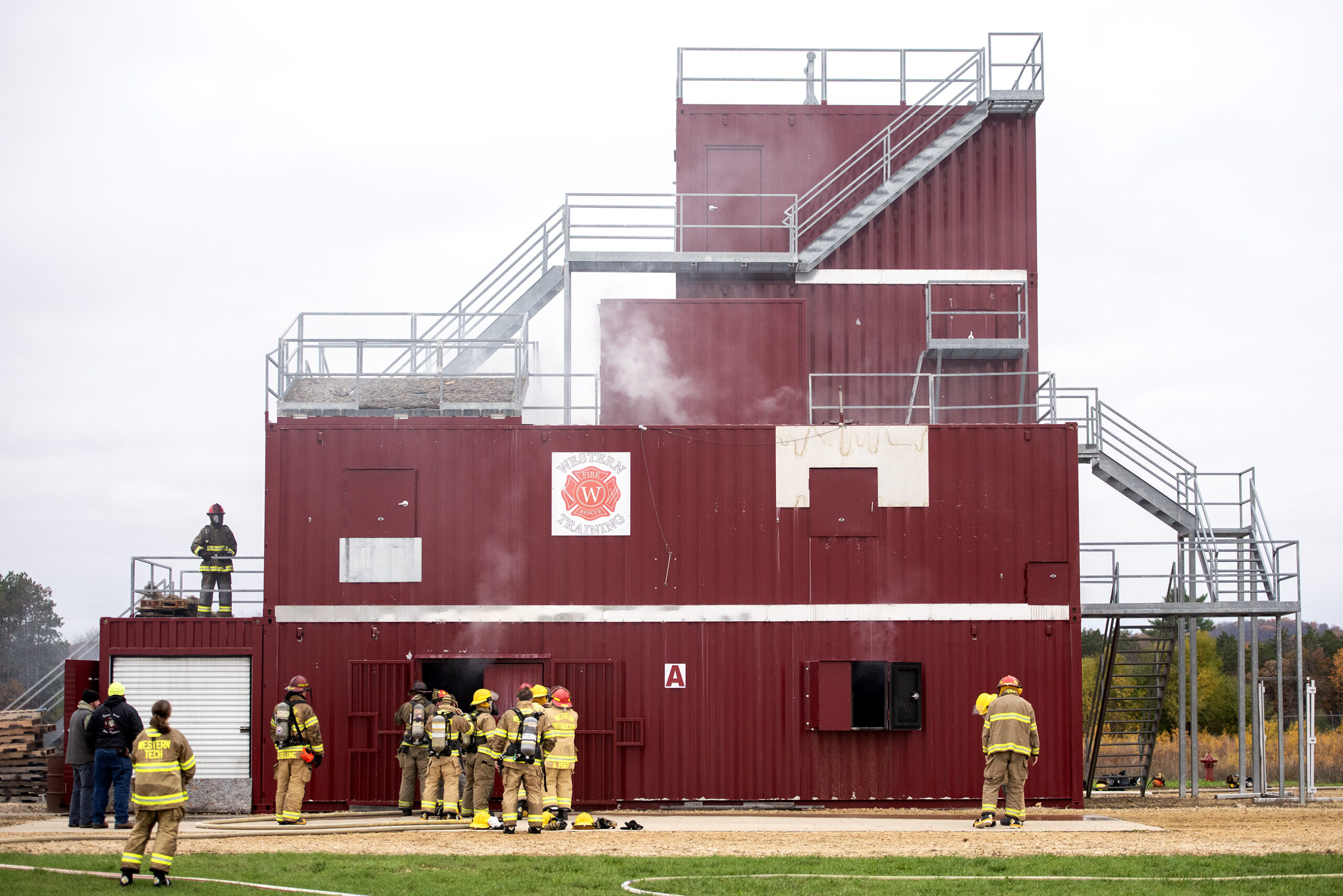 Smoke billows out of a red metal structure as participants in firefighter attire move around during the exercise.