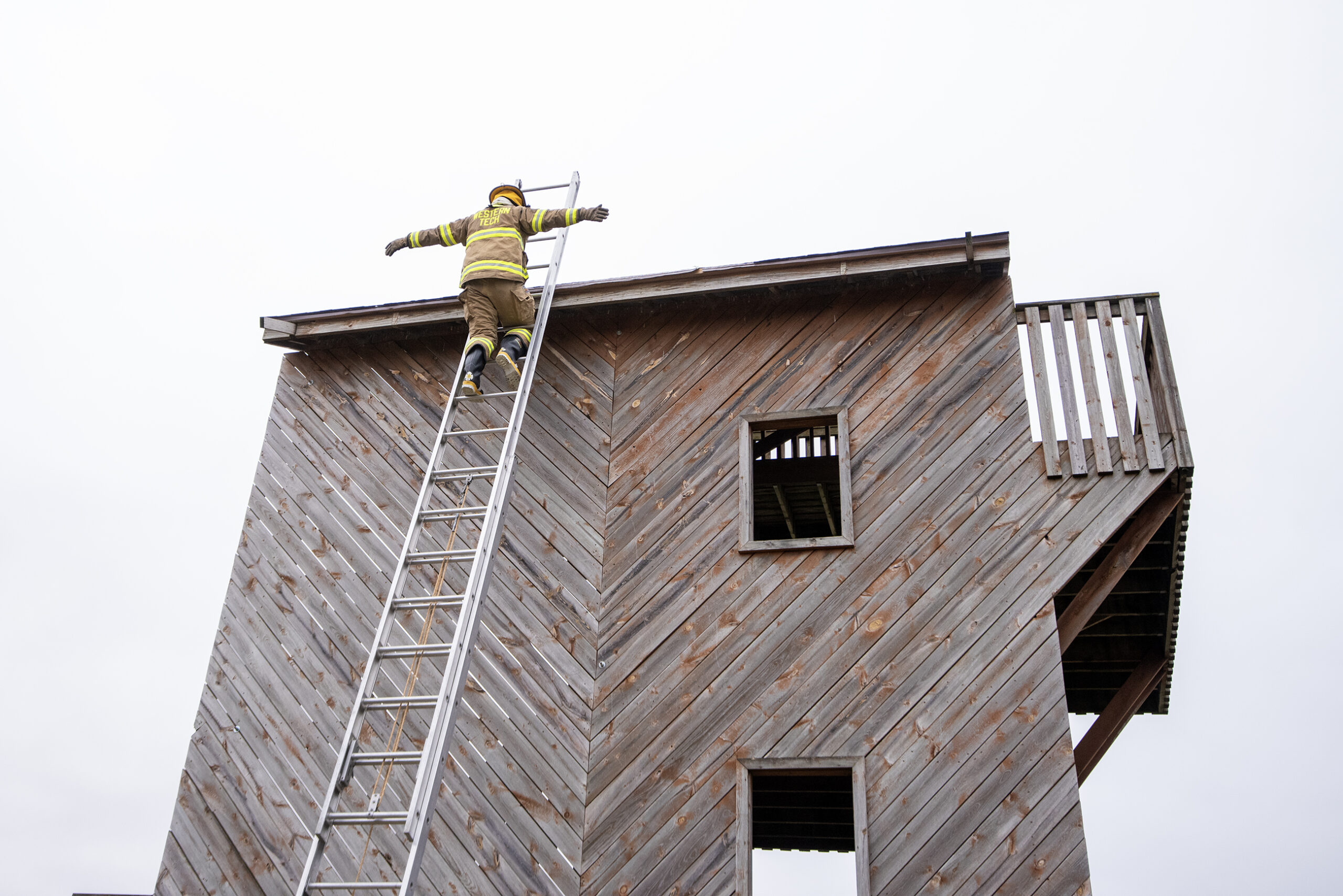 A student spreads their arms after reaching the roof of a small wooden structure by using a tall metal ladder.
