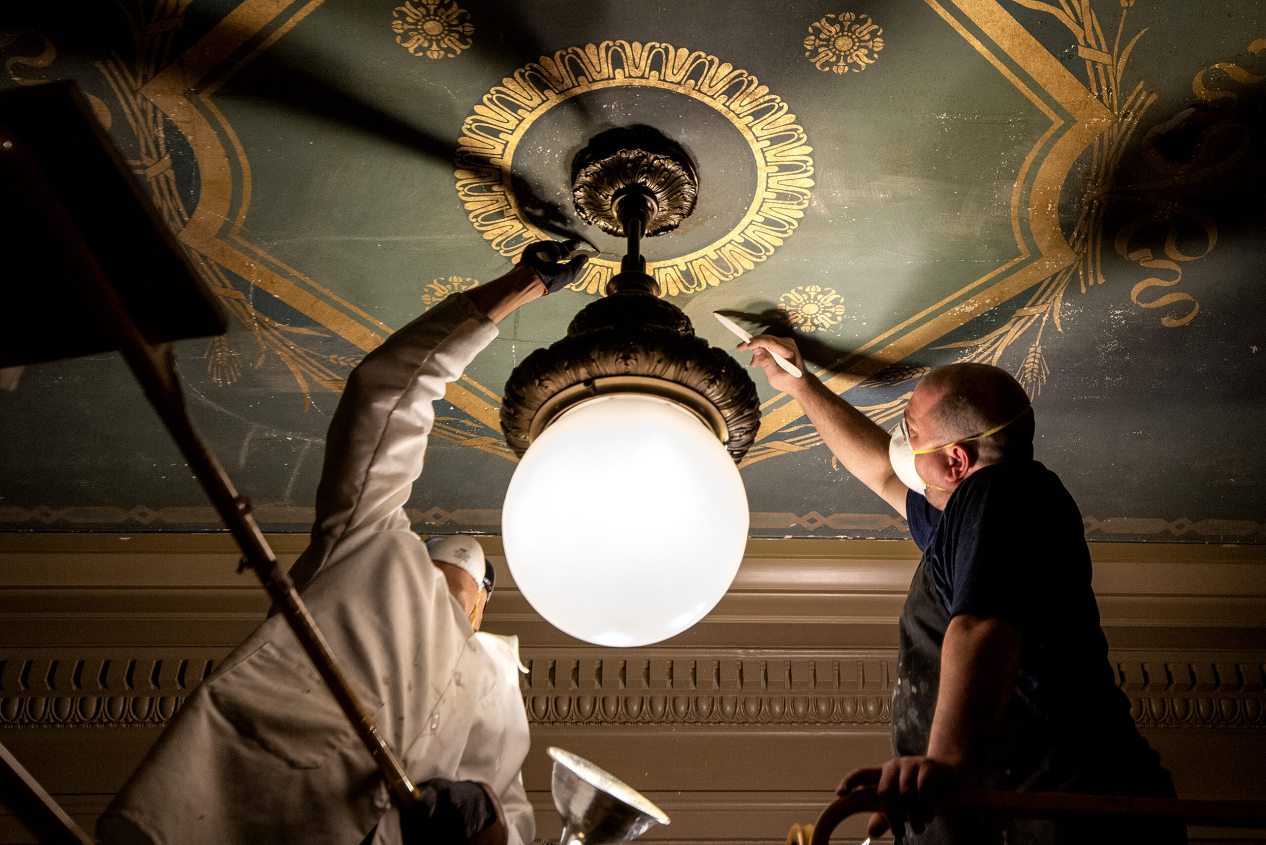 Two workers wear masks as they use a tool to scrape a decoratively painted ceiling.
