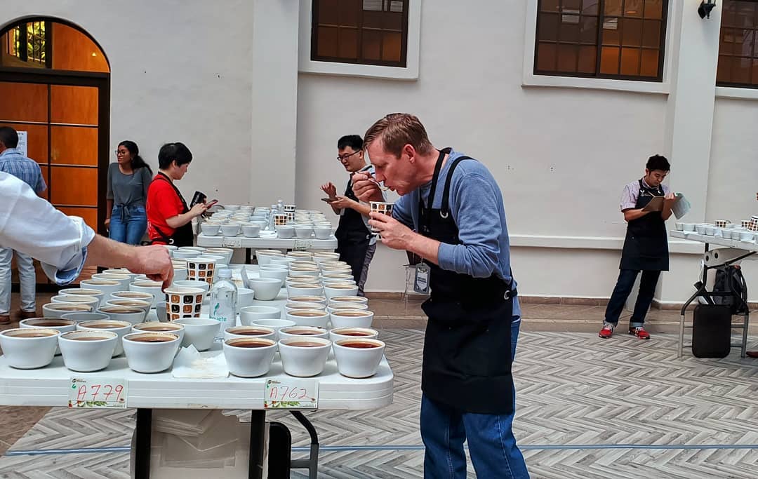 JBC Coffee Roasters evaluates some coffee as he stands in front of a table with many cups of coffee