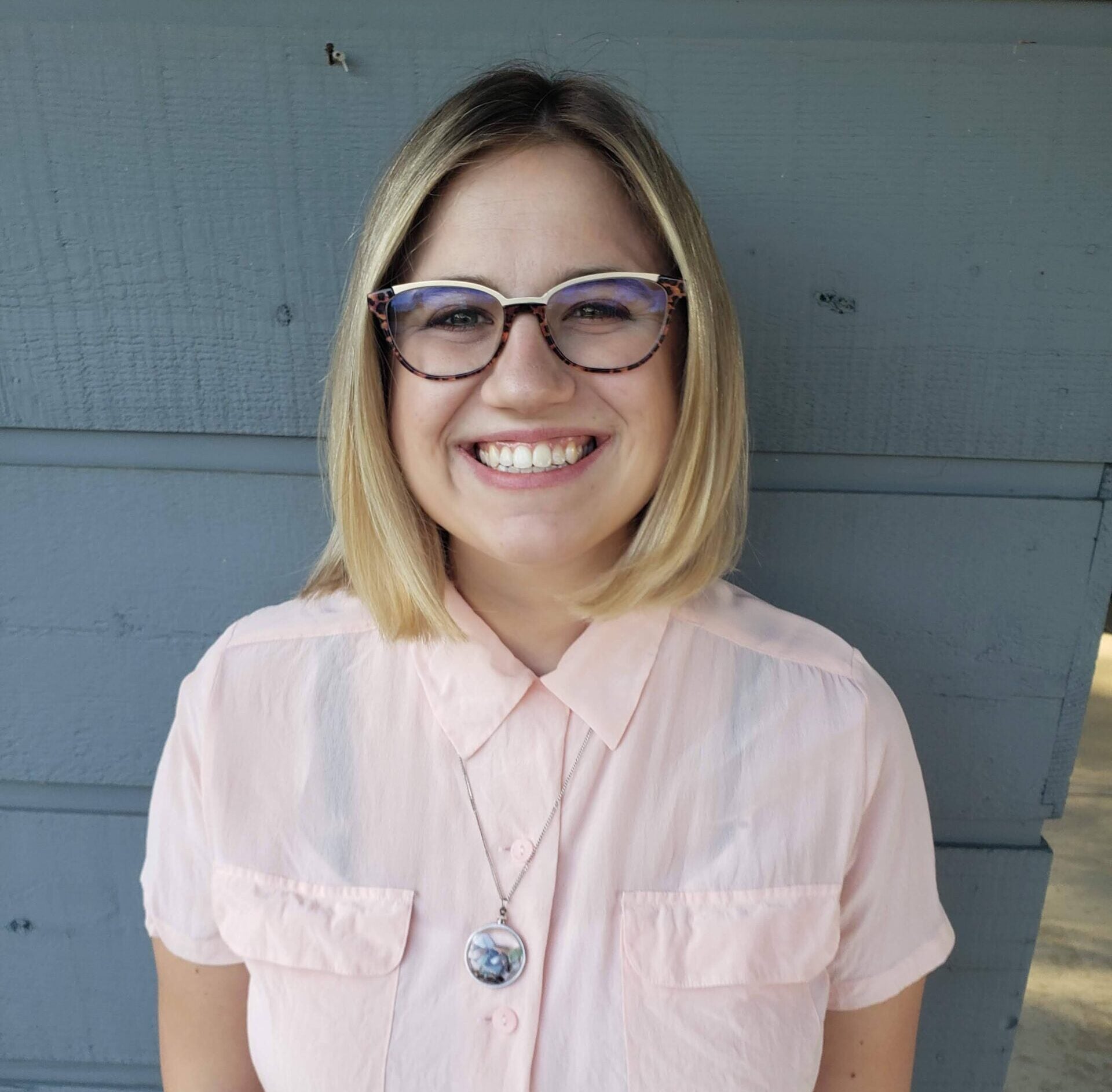 A person with chin-length blonde hair and glasses is smiling, wearing a light pink button-up shirt and a round pendant necklace, standing in front of a gray wooden wall.