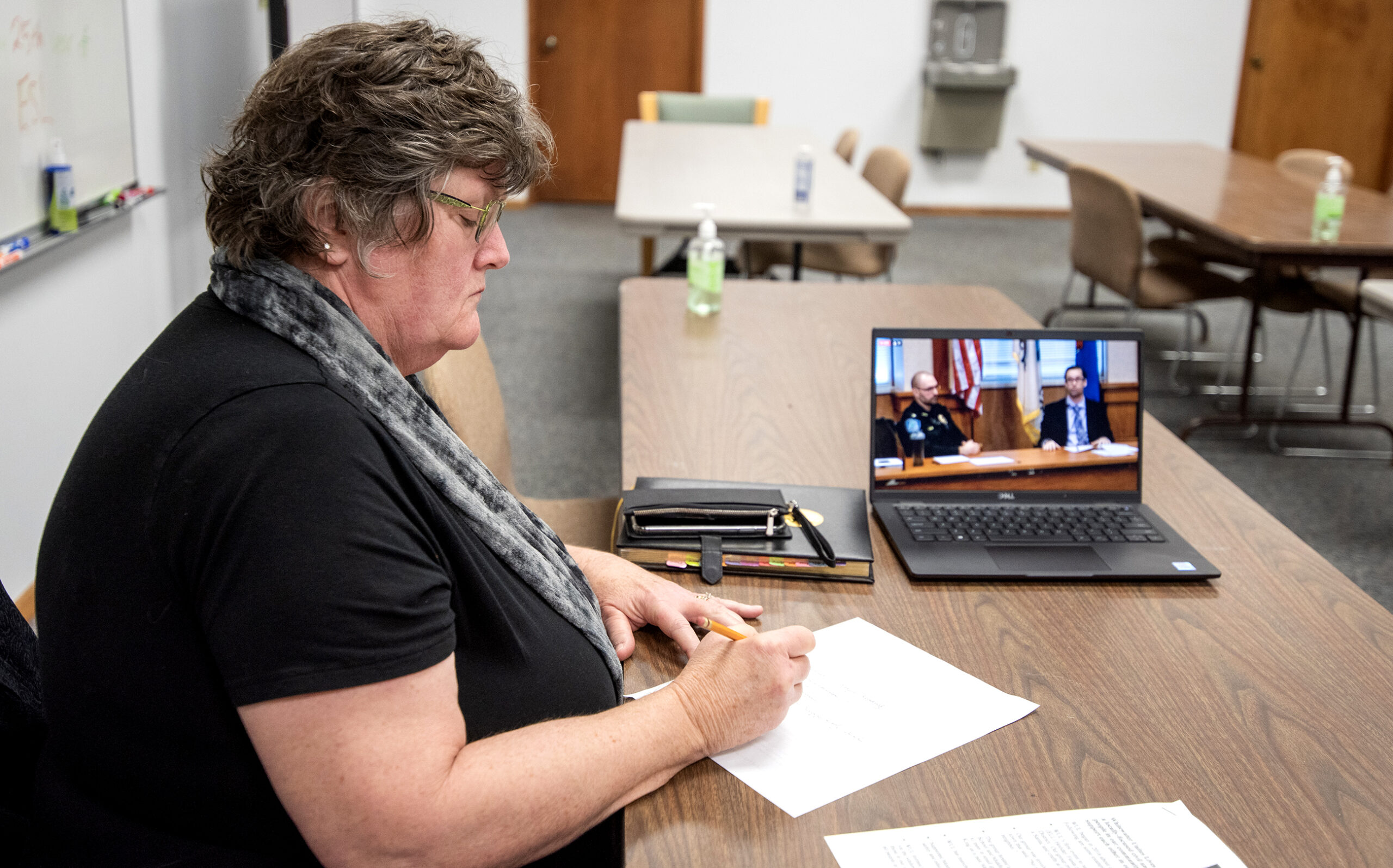 A woman takes notes as two men speak on a livestream shown on a laptop.