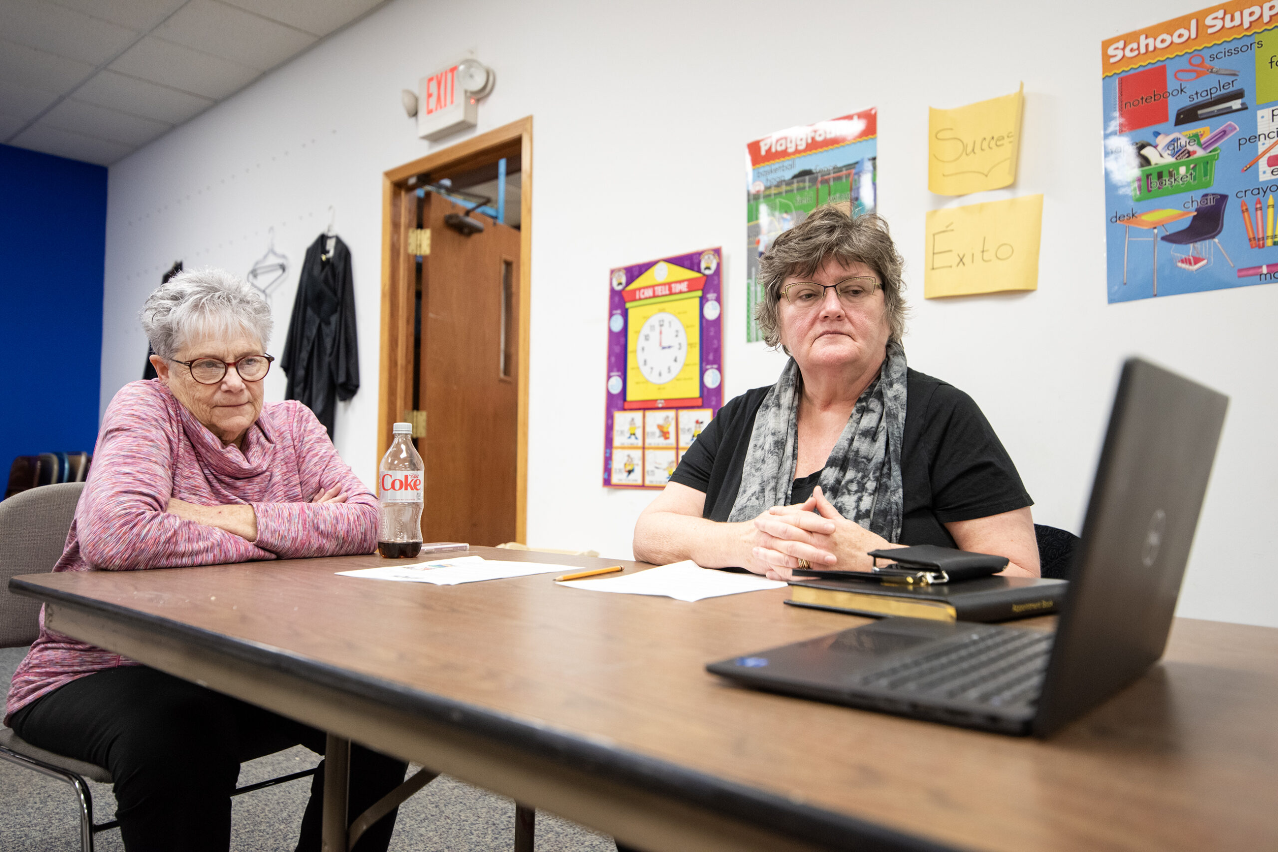 Two women sit at a table together watching a stream on a laptop.