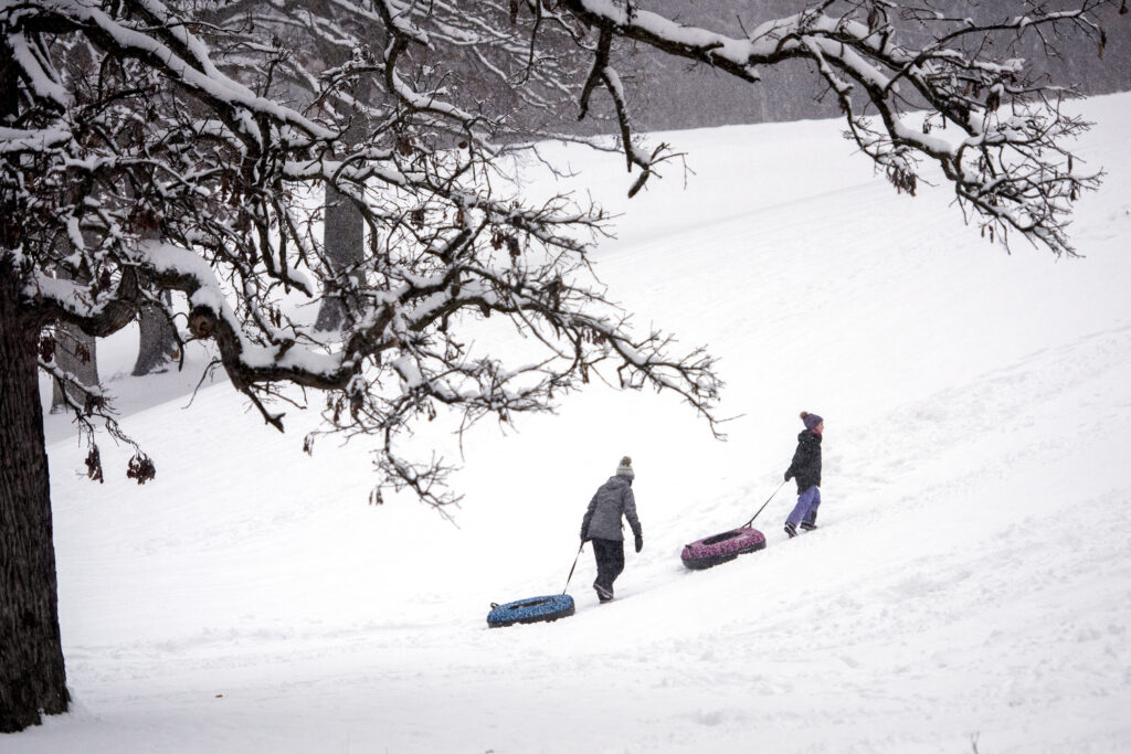 Two people pull inner tubes up a snowy hill. A snow-covered tree is in the foreground.