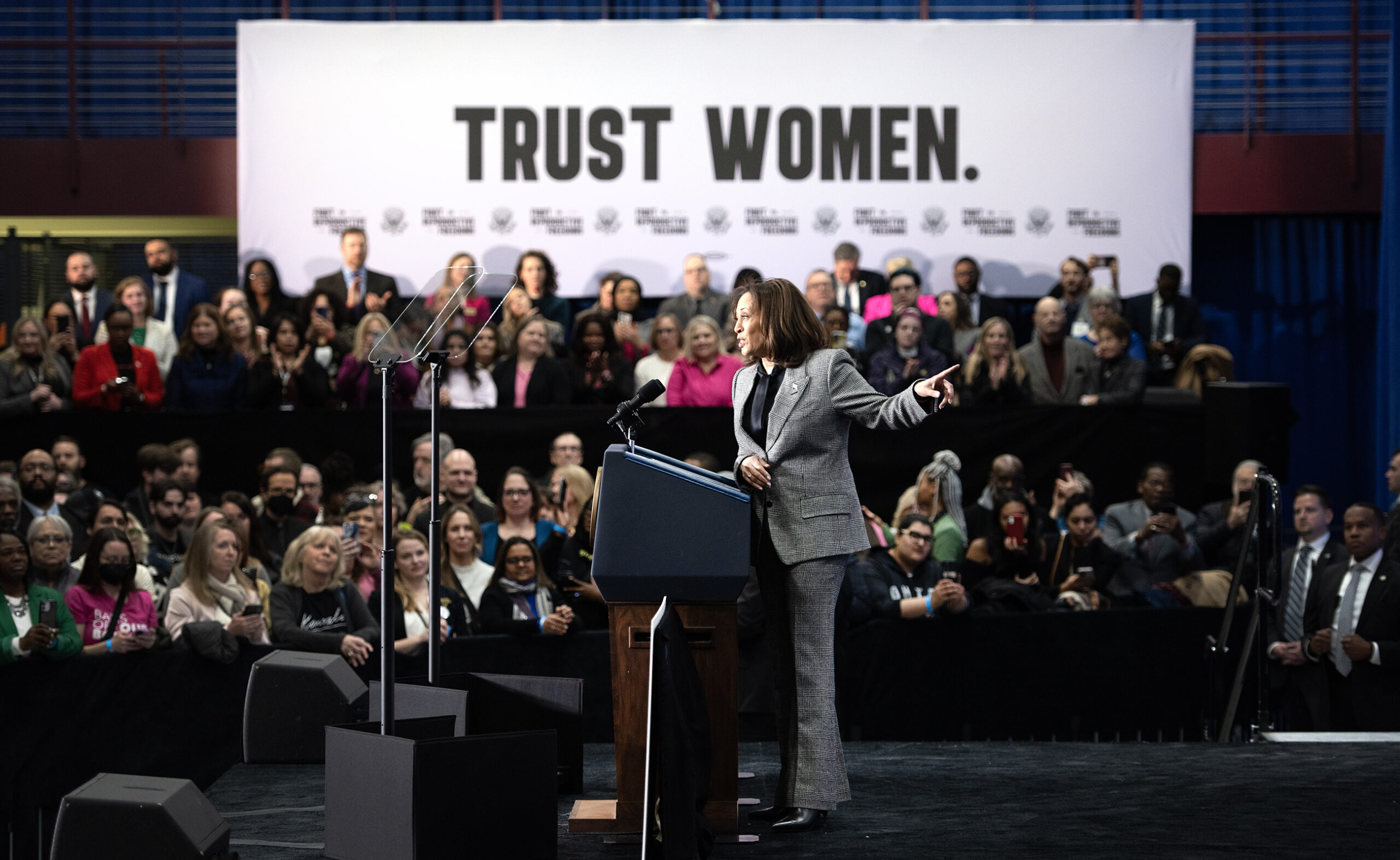 VP Harris points backward as she stands in front of a crowd & a banner that says "TRUST WOMEN."