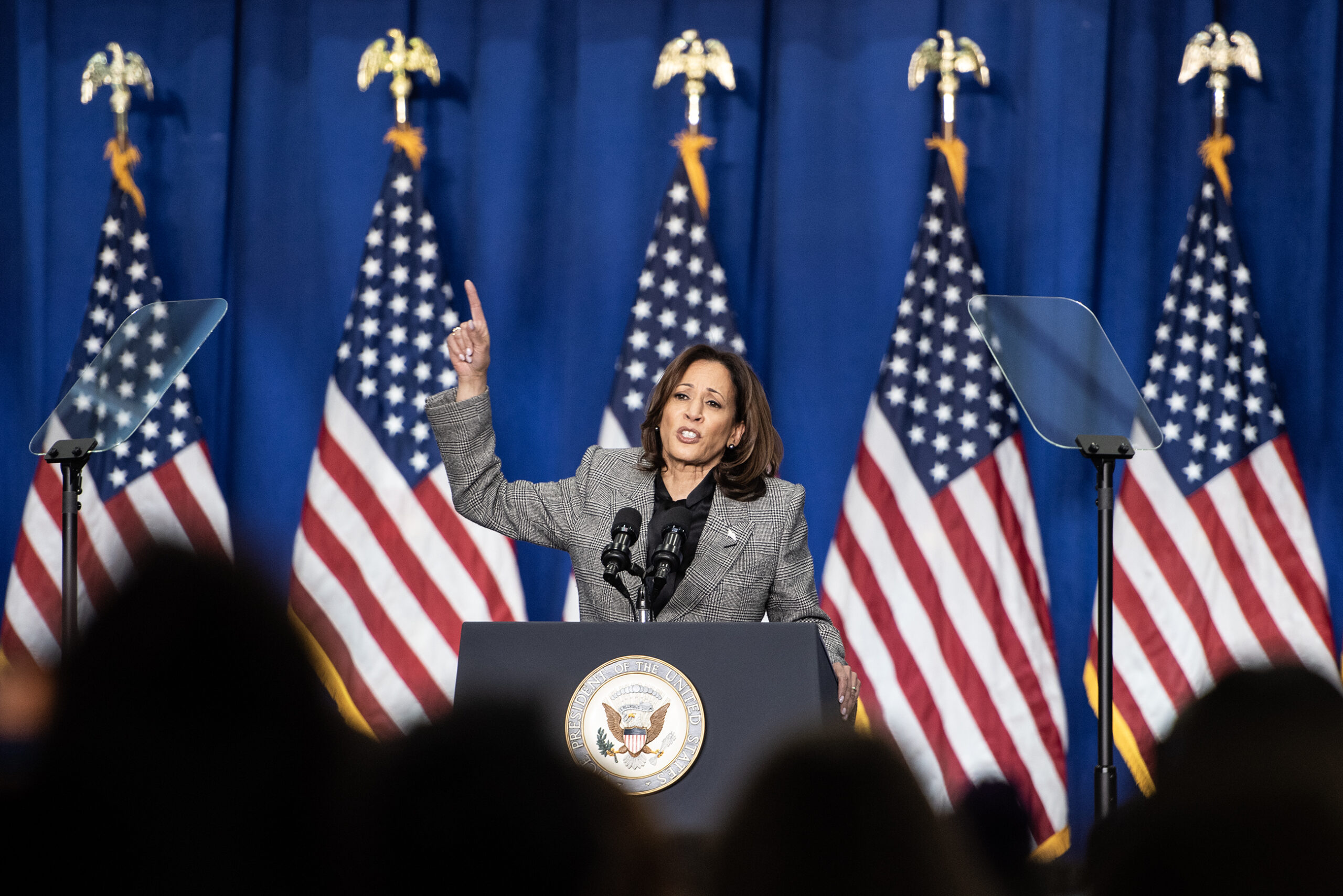 VP Harris points upward during a speech. Five US flags can be seen behind her.