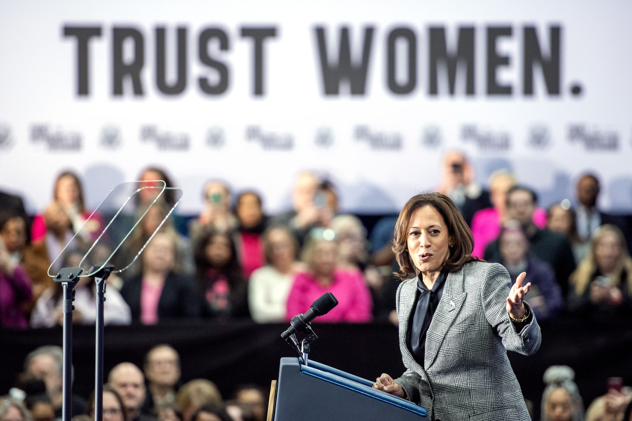 VP Harris gestures as she speaks at a podium. The words "TRUST WOMEN" appear behind her on a black and white banner.