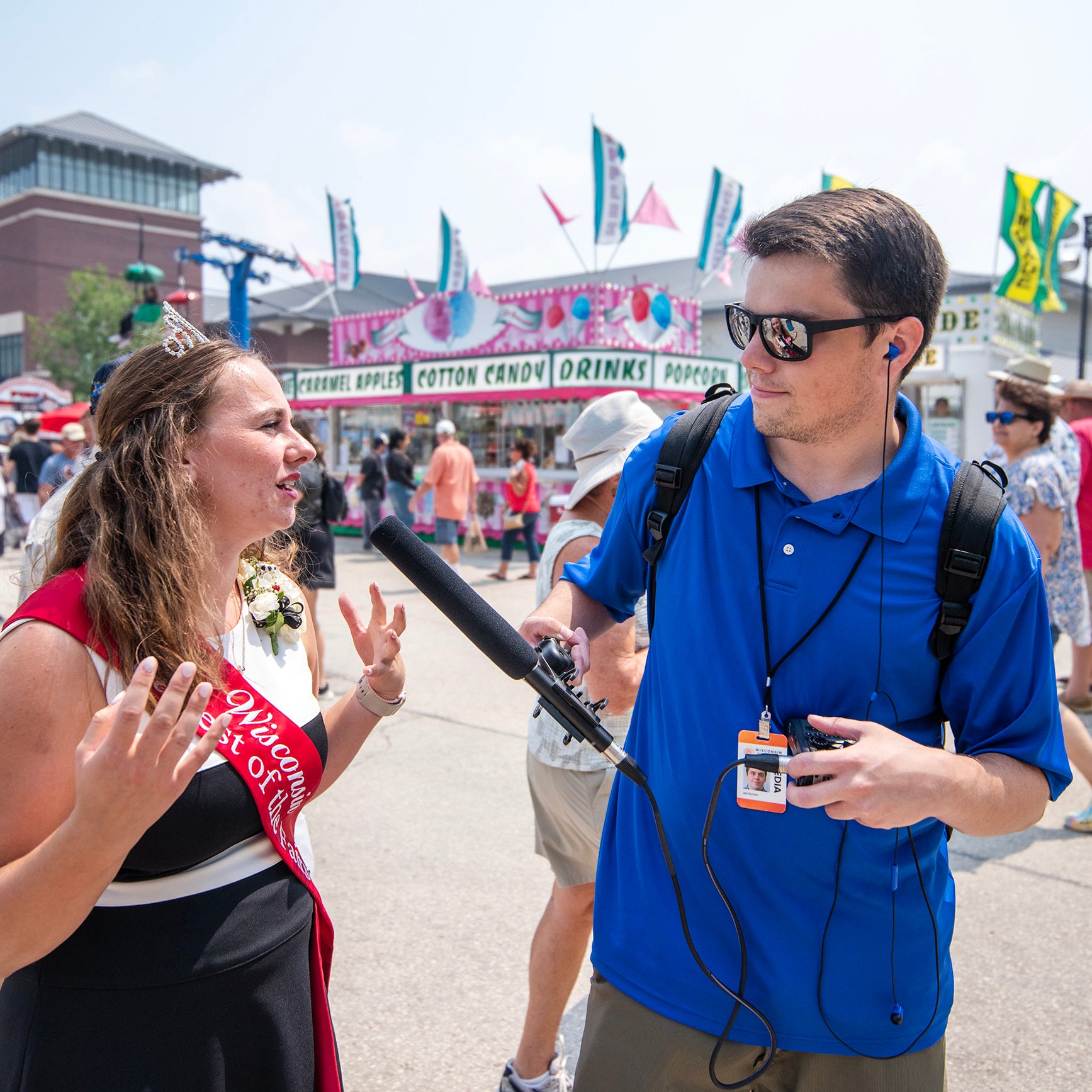 A woman wearing a sash and crown is interviewed by a man holding a microphone at an outdoor fair with food stands in the background.