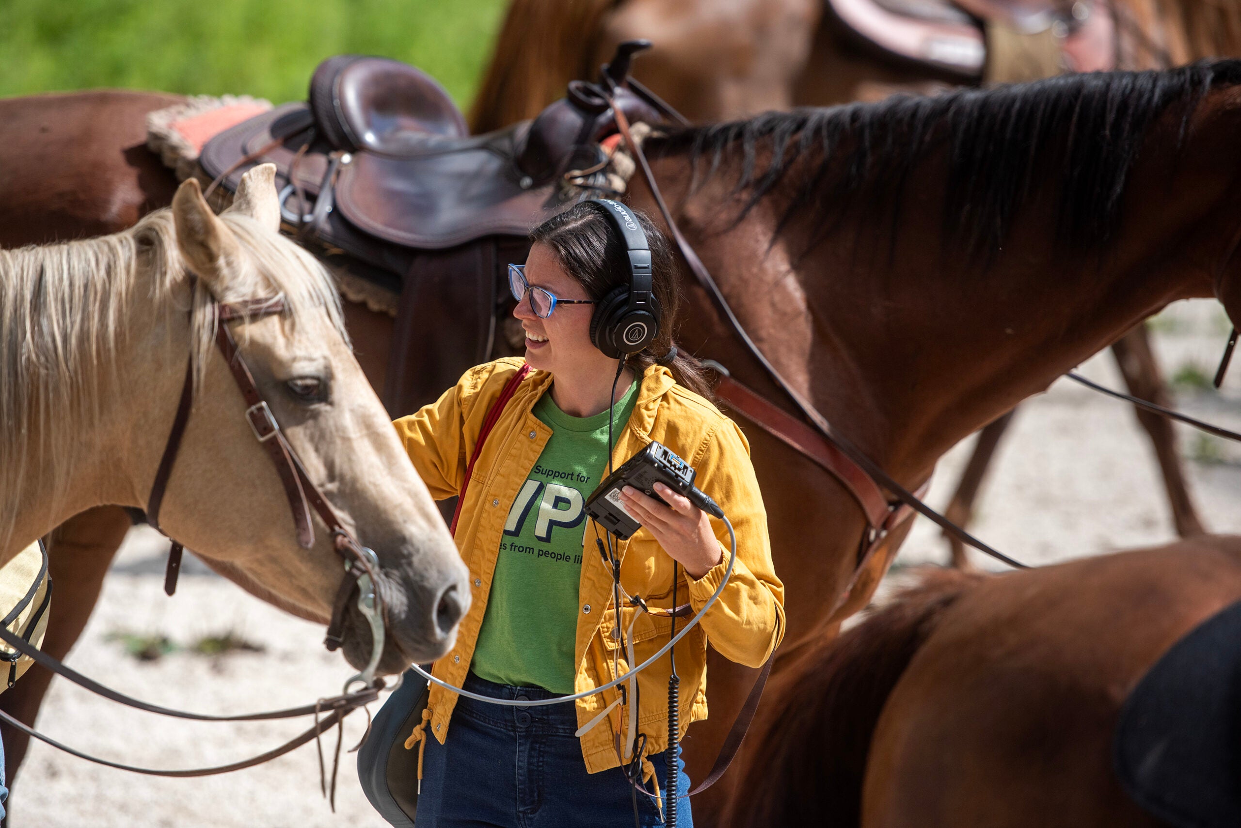 A person wearing headphones and holding audio equipment stands next to a light-colored horse, with other horses and saddles visible in the background.