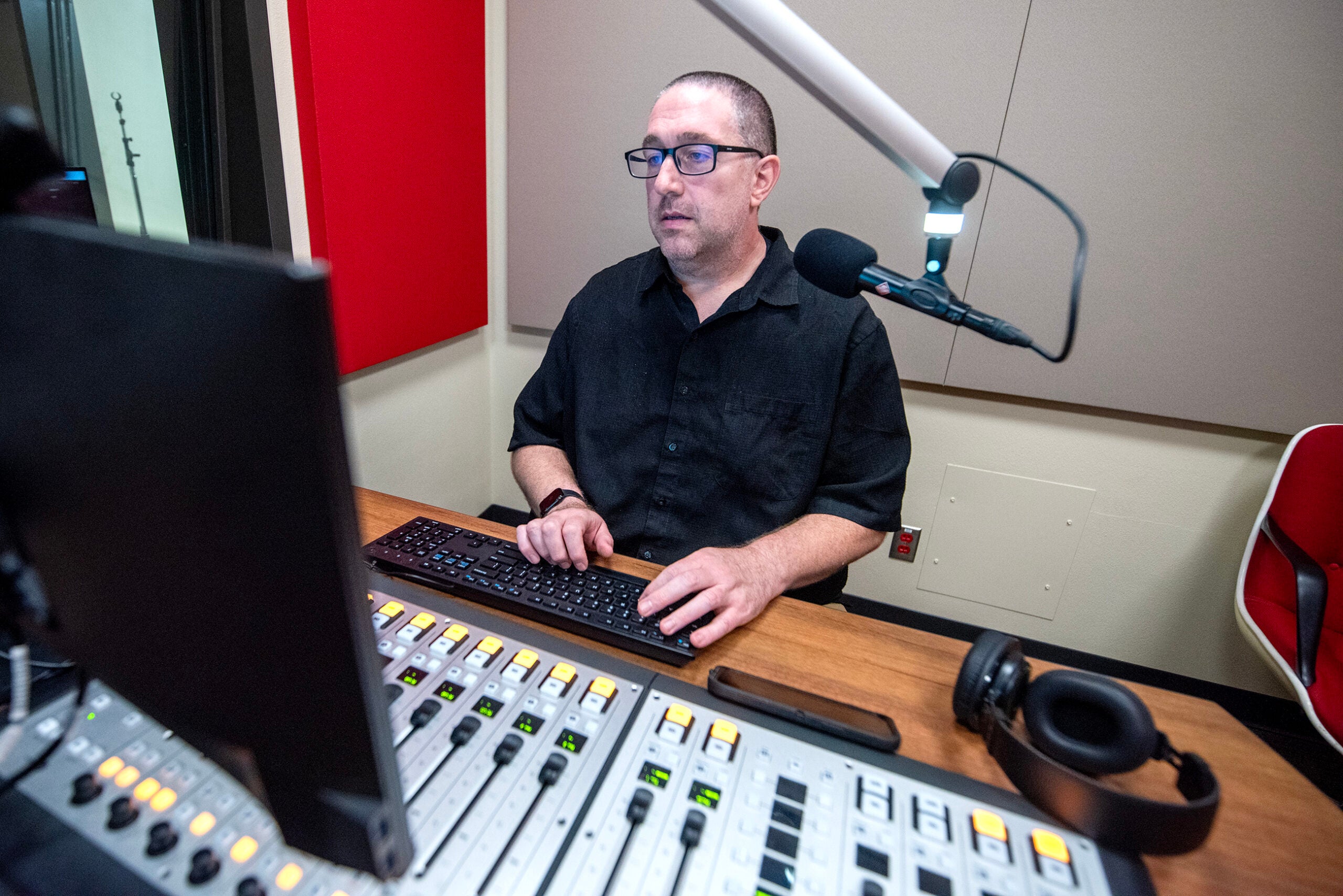 A man sits at a radio studio desk using a computer keyboard, with a microphone, audio mixer, monitor, and headphones in view.