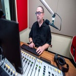 A man sits at a radio studio desk using a computer keyboard, with a microphone, audio mixer, monitor, and headphones in view.
