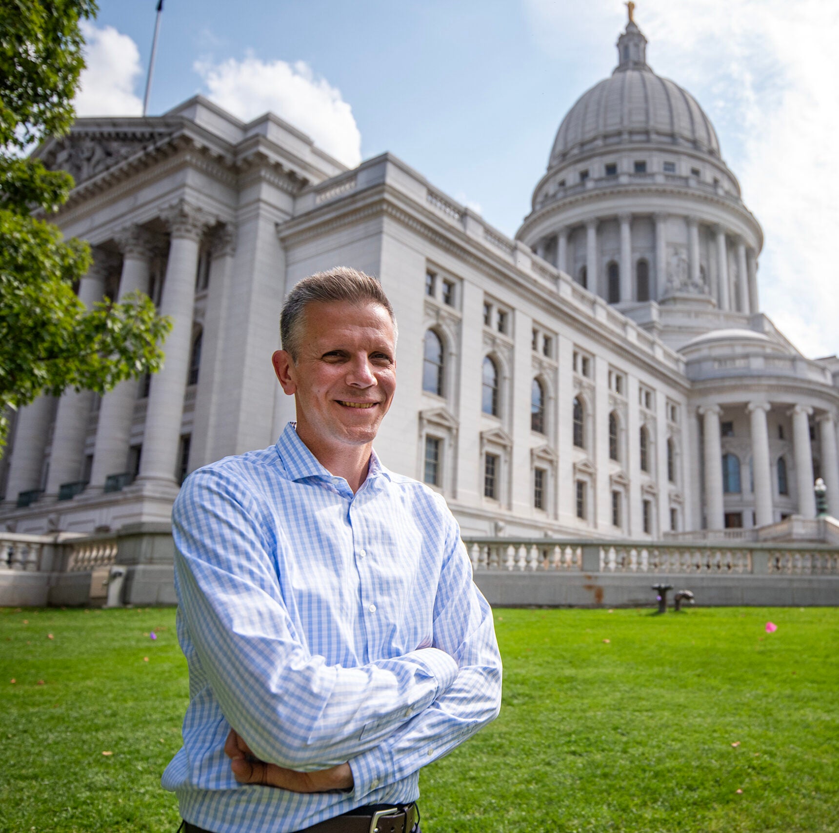 A man in a light blue striped shirt stands with folded arms on the lawn in front of a large government building with a dome.