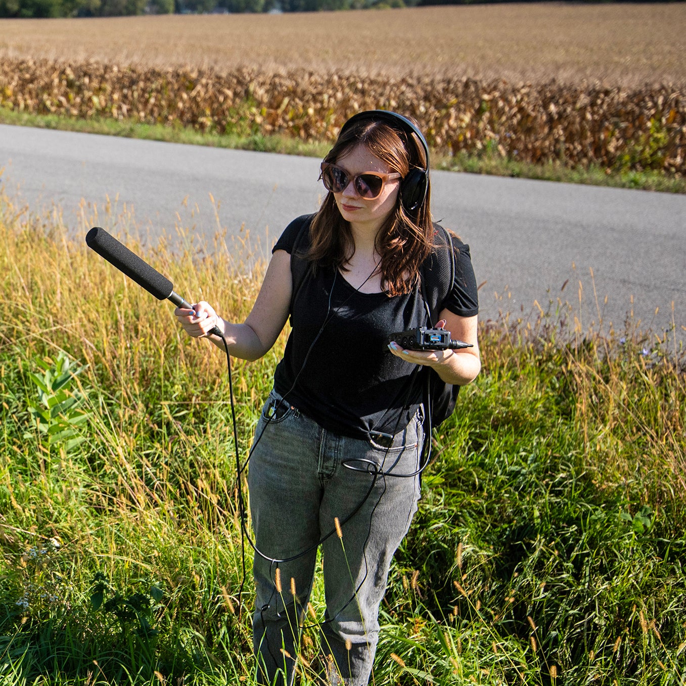 A woman with headphones holds a microphone and audio recorder, standing in tall grass by a rural roadside on a sunny day.