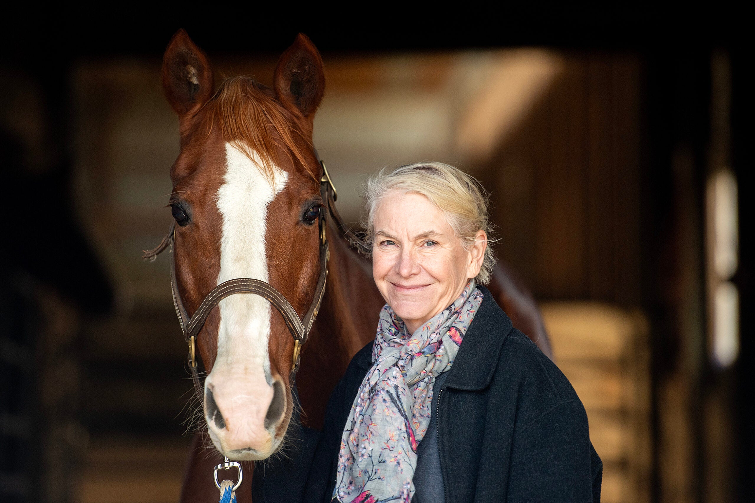 A woman with light hair stands next to a brown horse with a white blaze, both facing the camera, inside a barn.