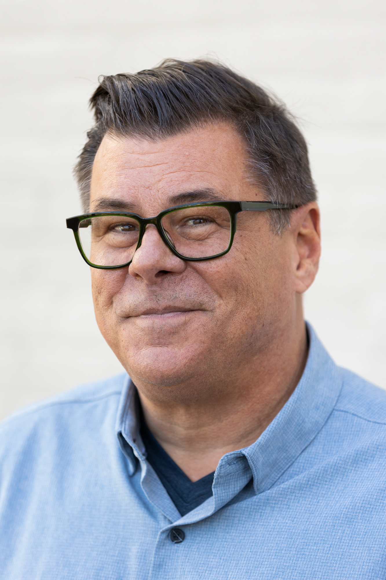 A middle-aged man with dark hair and glasses, wearing a light blue shirt, smiles slightly while standing in front of a light-colored background.