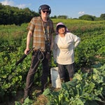 A man with audio equipment stands next to a woman in a sunhat on a vegetable farm, surrounded by green plants under a blue sky.
