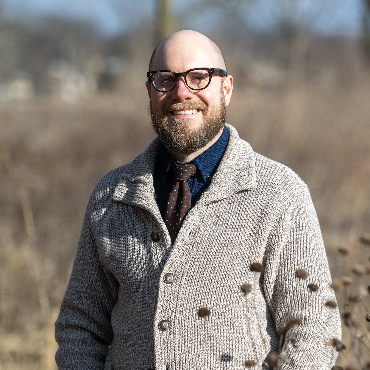 A bearded man wearing glasses, a gray knit sweater, and a polka-dot tie stands outdoors in a sunny, dry field.