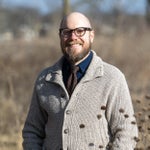 A bearded man wearing glasses, a gray knit sweater, and a polka-dot tie stands outdoors in a sunny, dry field.