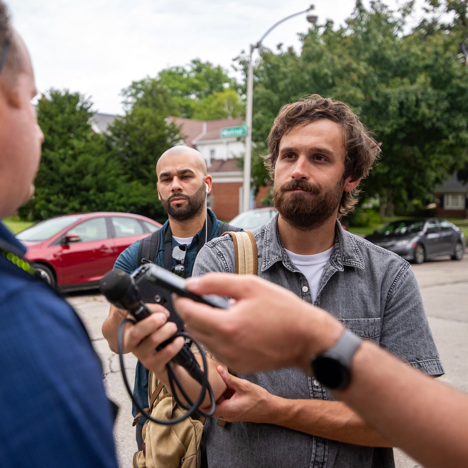Two men stand on a residential street, one holding audio recording equipment toward a person out of frame; cars and trees are visible in the background.