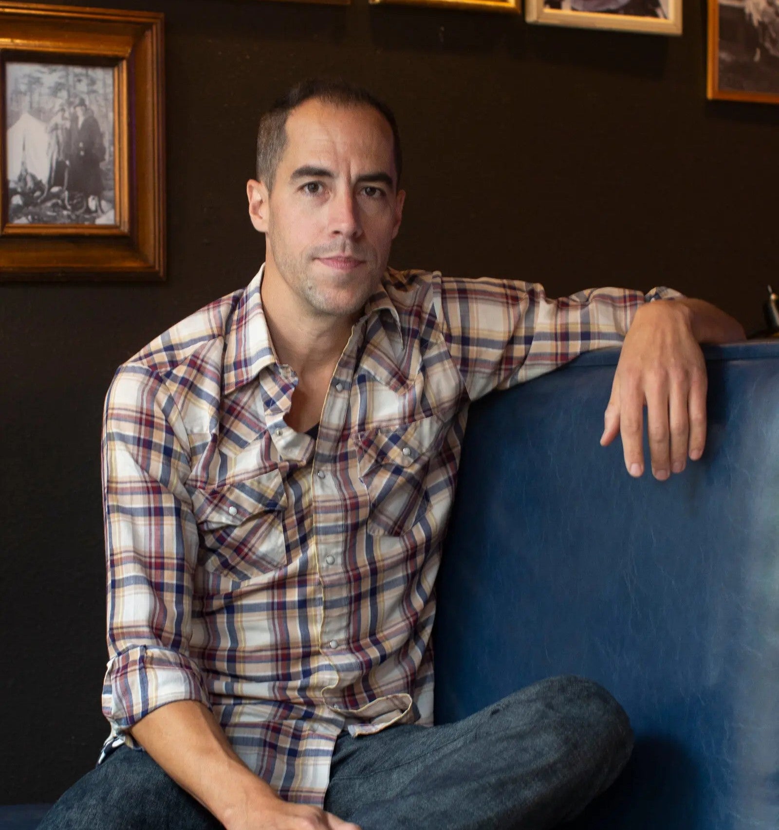 A man in a plaid shirt sits on a blue bench with one arm resting on the back, looking at the camera. Framed photos hang on the dark wall behind him.