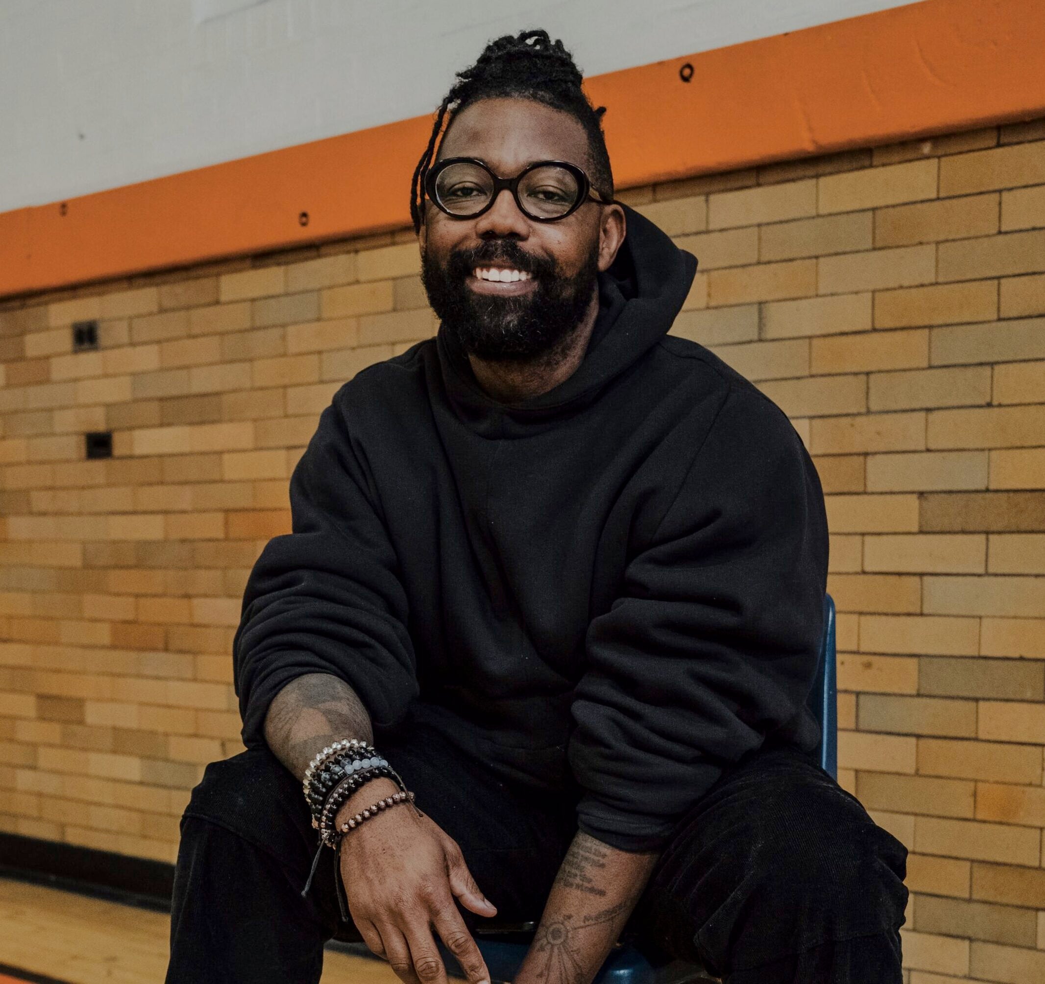 A man with glasses and a beard sits on a chair in a gymnasium, wearing a black hoodie, black pants, white socks, and black shoes.
