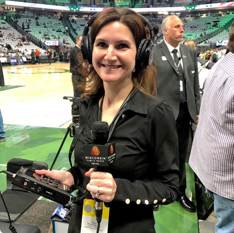 A woman wearing headphones holds a microphone and audio recorder courtside at a basketball arena, with people and empty seats in the background.