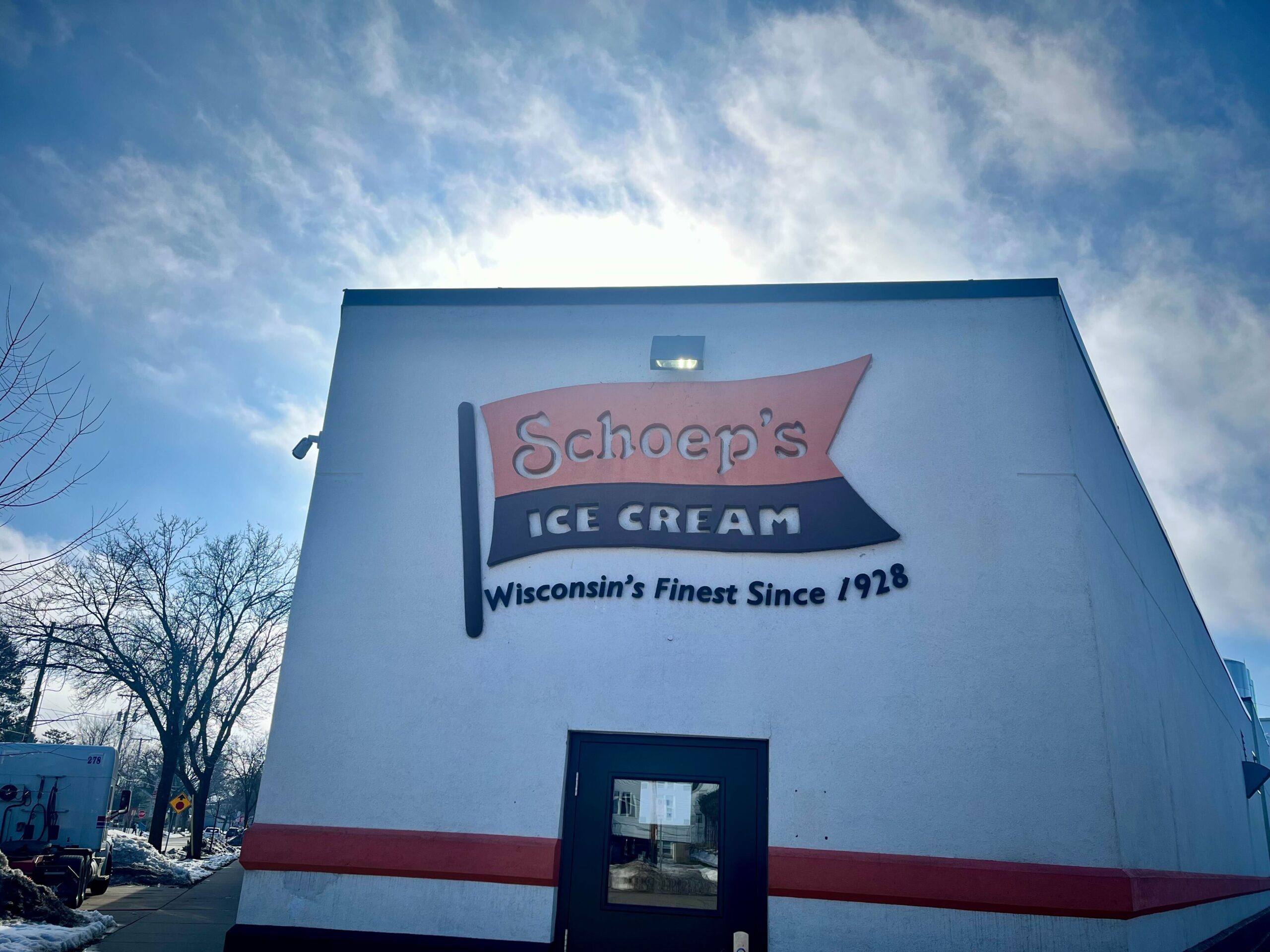 The front of Schoep's ice cream factory -- an orange and brown sign on a white building, against a sunny winter sky.