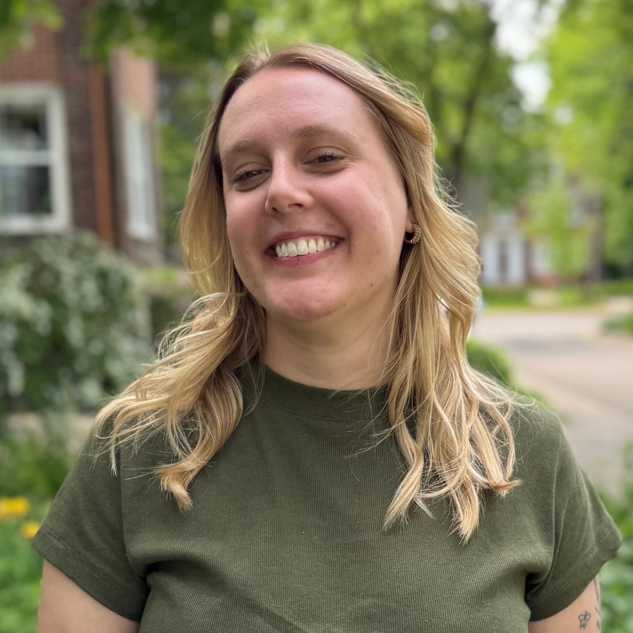 A woman with long blonde hair and a green t-shirt smiles outdoors, standing in front of greenery and a blurred building background.