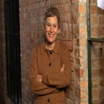 A person with short hair and a brown coat stands indoors against a brick wall, arms crossed, and smiling at the camera.