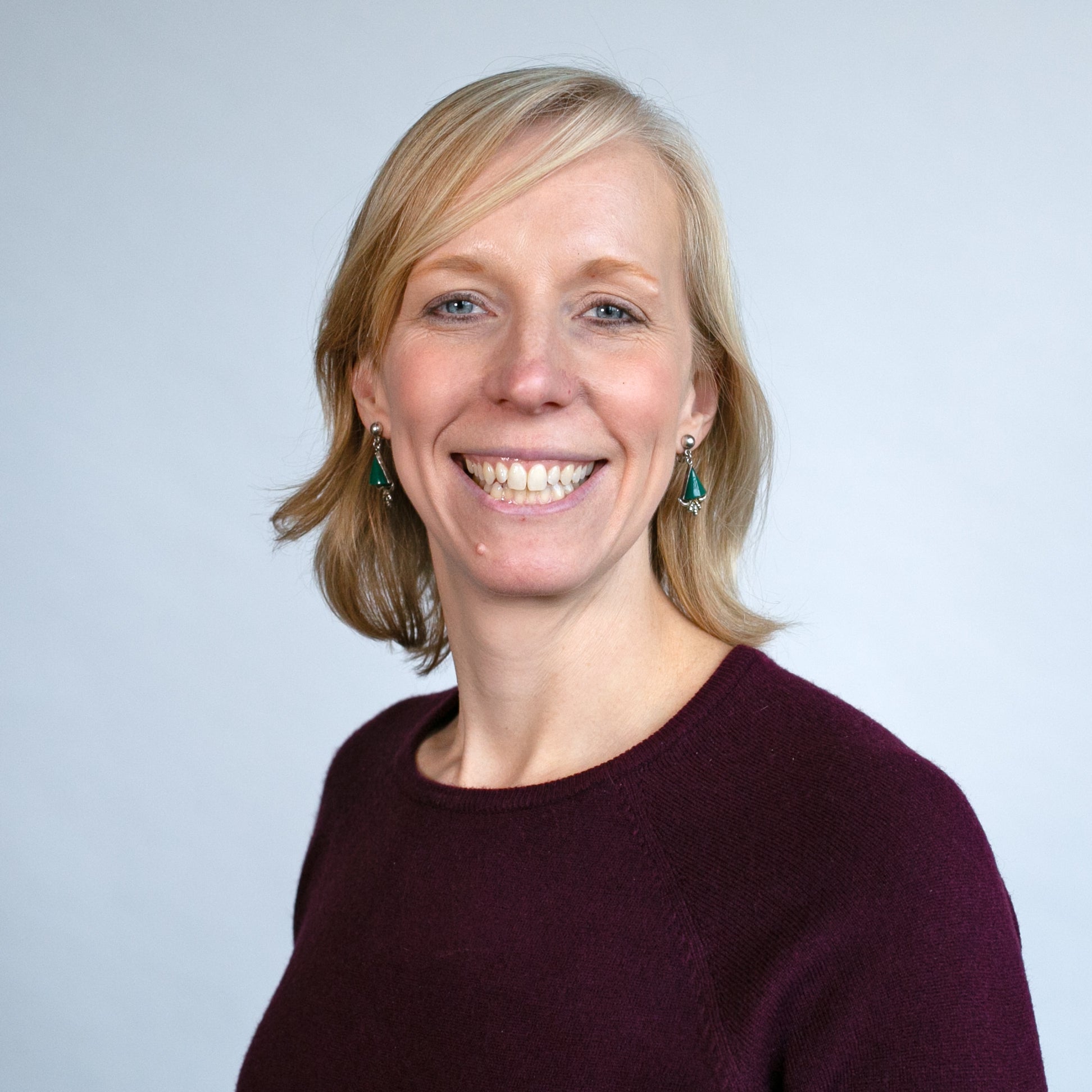 A woman with blonde hair and green earrings smiles at the camera, wearing a burgundy sweater against a plain light background.