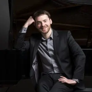 A man in a suit sits casually with one arm resting on a piano and the other on his knee, smiling at the camera.